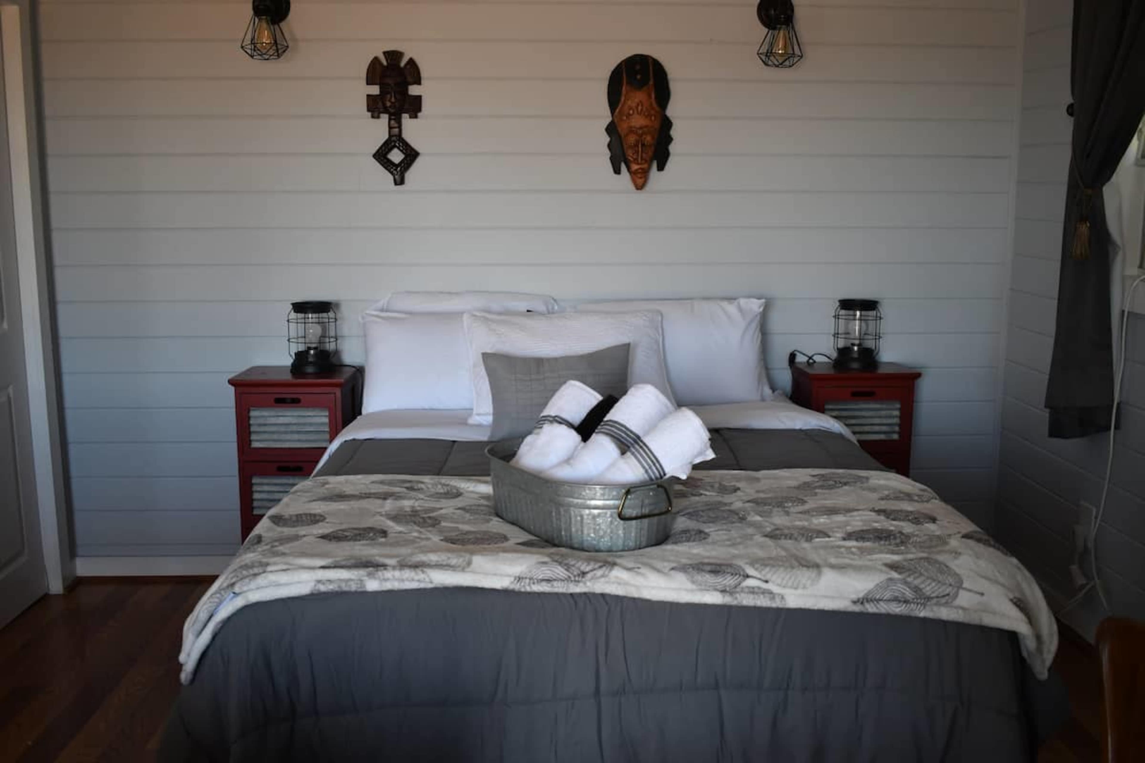 A rustic bedroom featuring a bed with gray and white linens, a decorative throw blanket, and a tray of rolled towels, framed by wooden paneling and wall-mounted masks.
