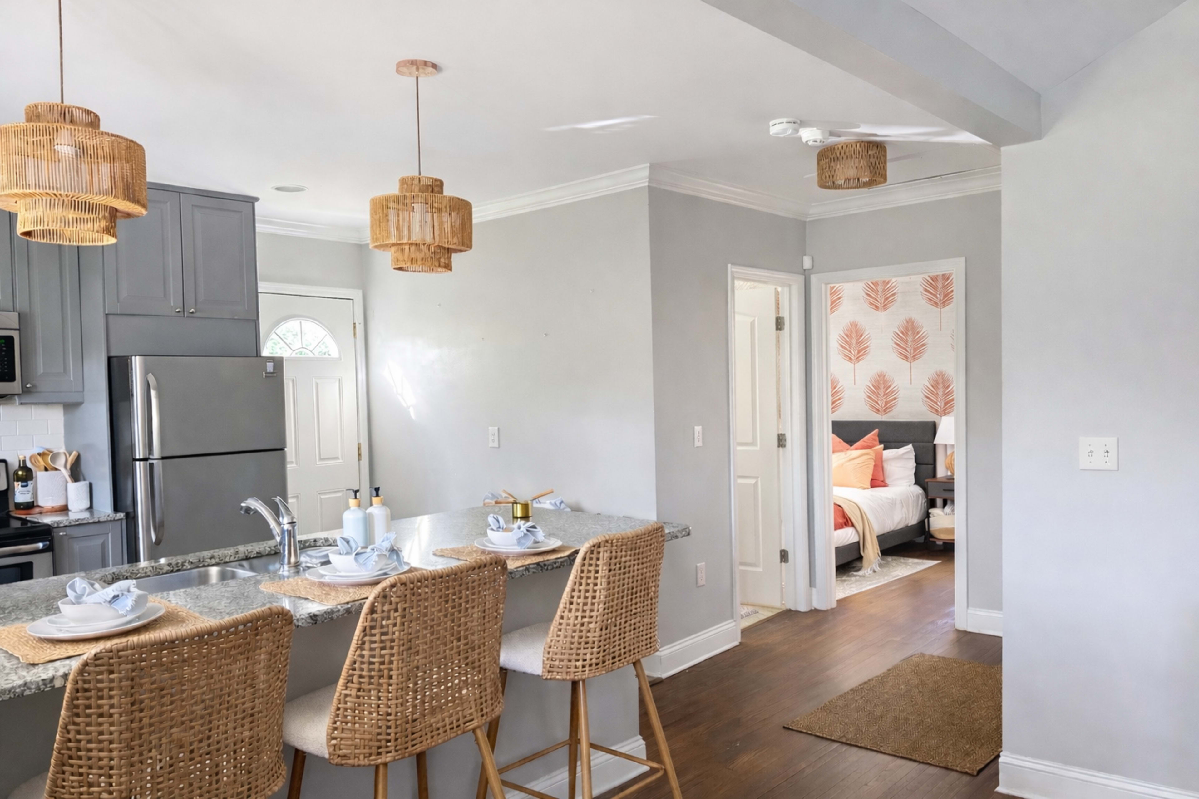 The image shows a modern kitchen with gray cabinets, woven bar stools at a granite countertop, and a door leading to a well-decorated living space featuring a bed and decorative wall art.