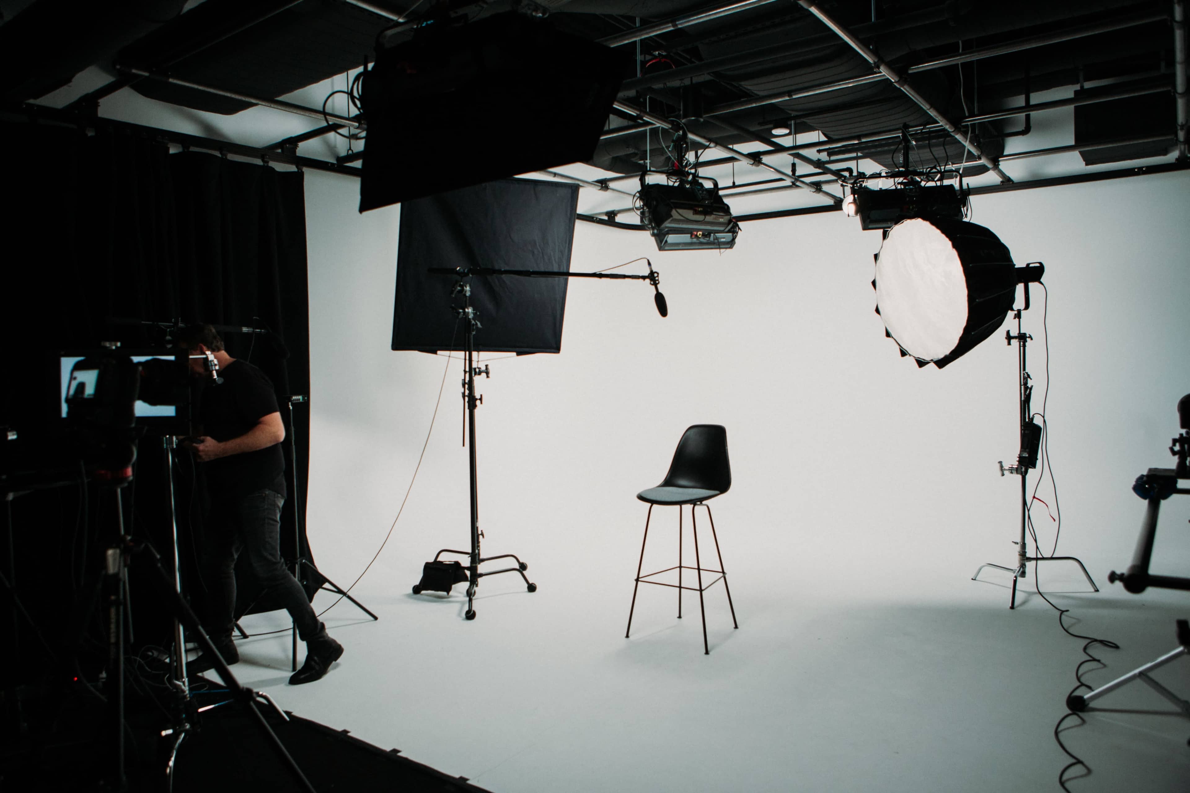 A film studio setup features a bare white backdrop, a high stool, and various lighting equipment.