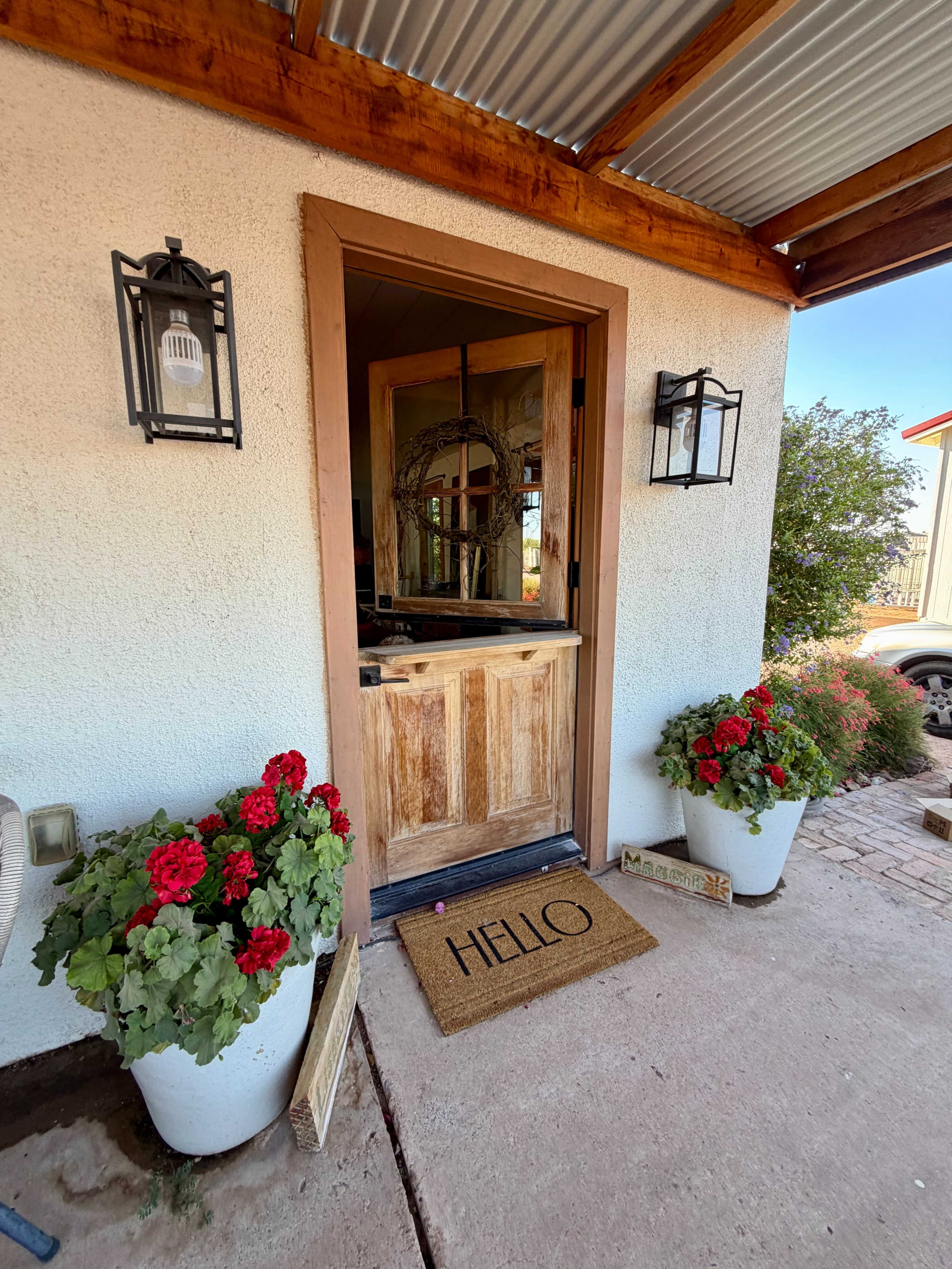 A wooden front door with a "HELLO" doormat is framed by potted flowers and outdoor lanterns.