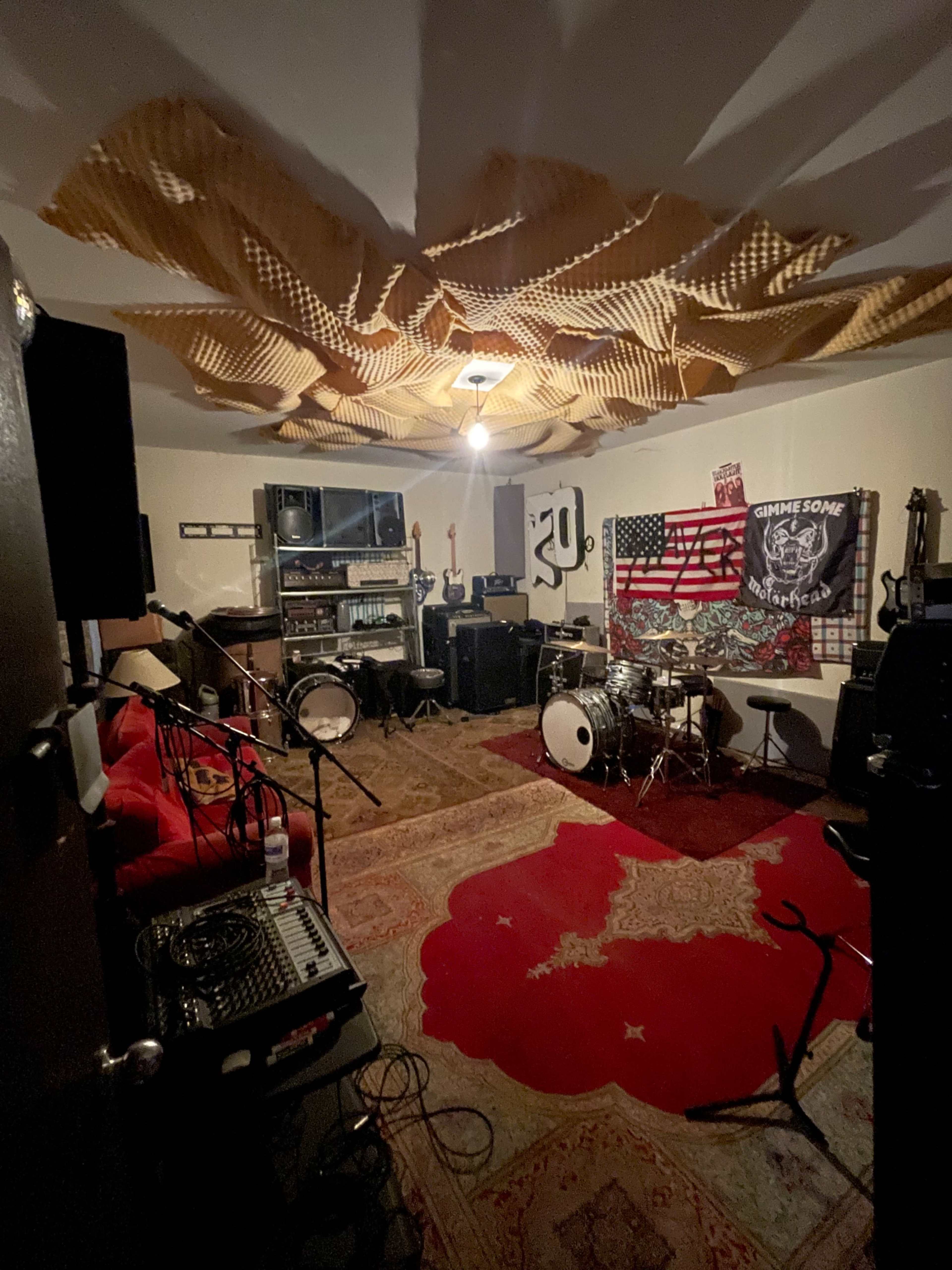 A music rehearsal room is filled with instruments, amplifiers, and soundproofing materials, featuring a drum set, guitars, and various flags on the walls.