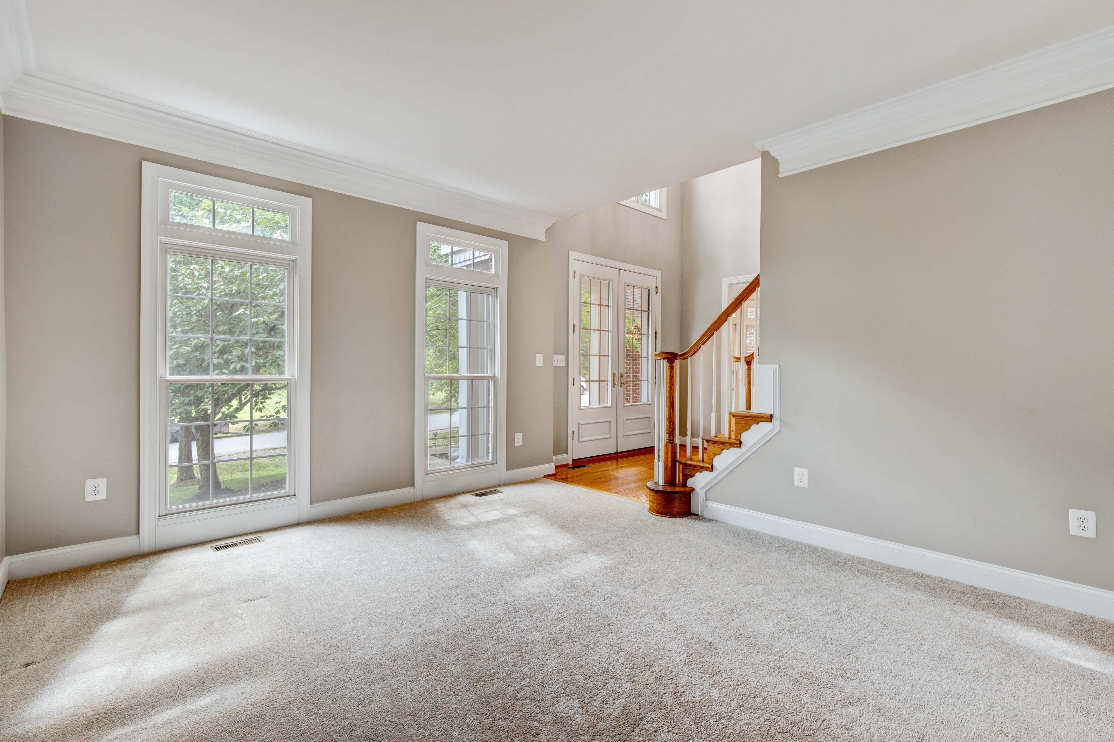 An empty living room with large windows, a staircase in the corner, and light-colored walls.
