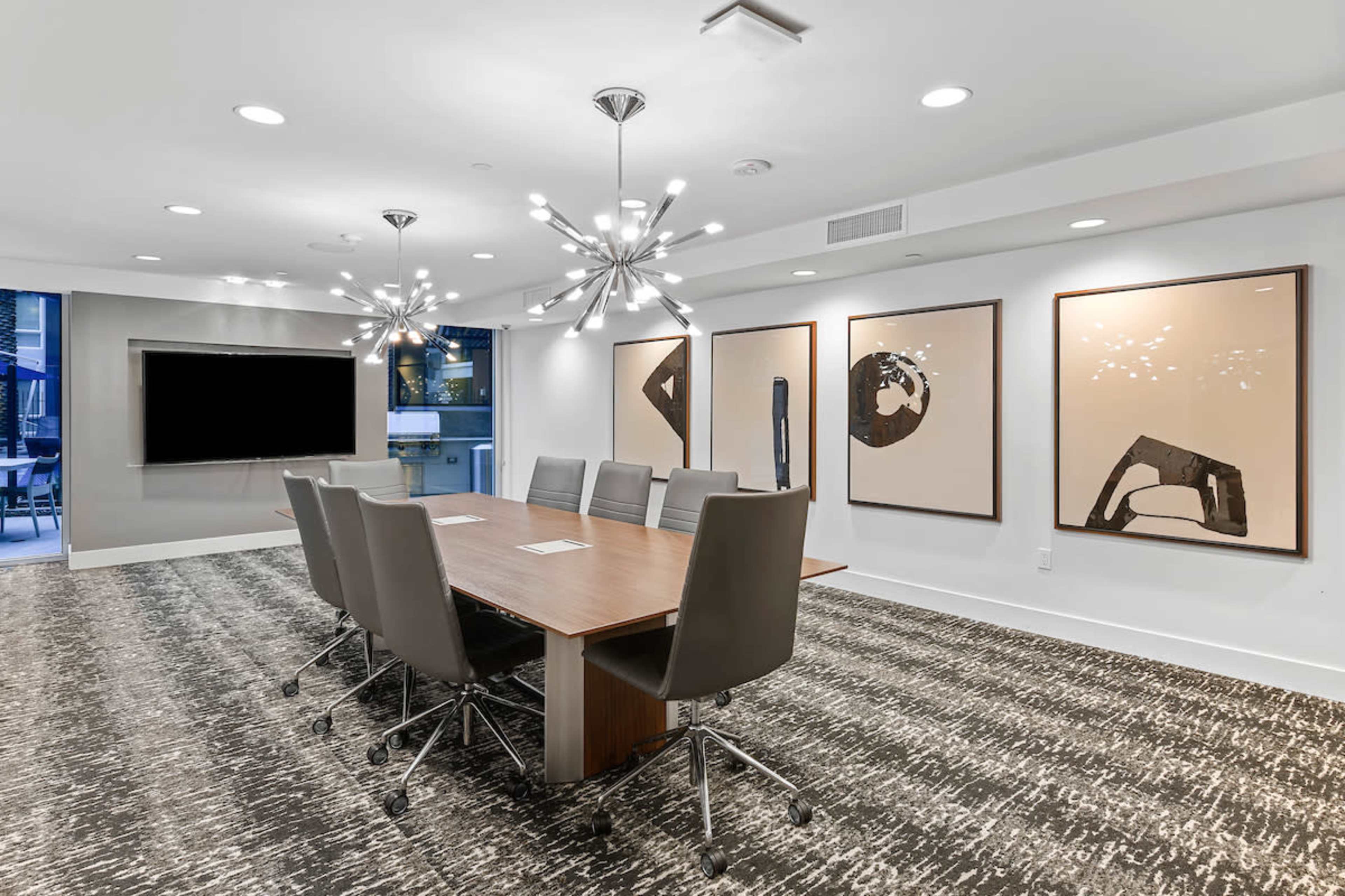 A modern conference room features a long wooden table surrounded by ergonomic chairs, with abstract artwork hanging on the walls and a large screen mounted for presentations.