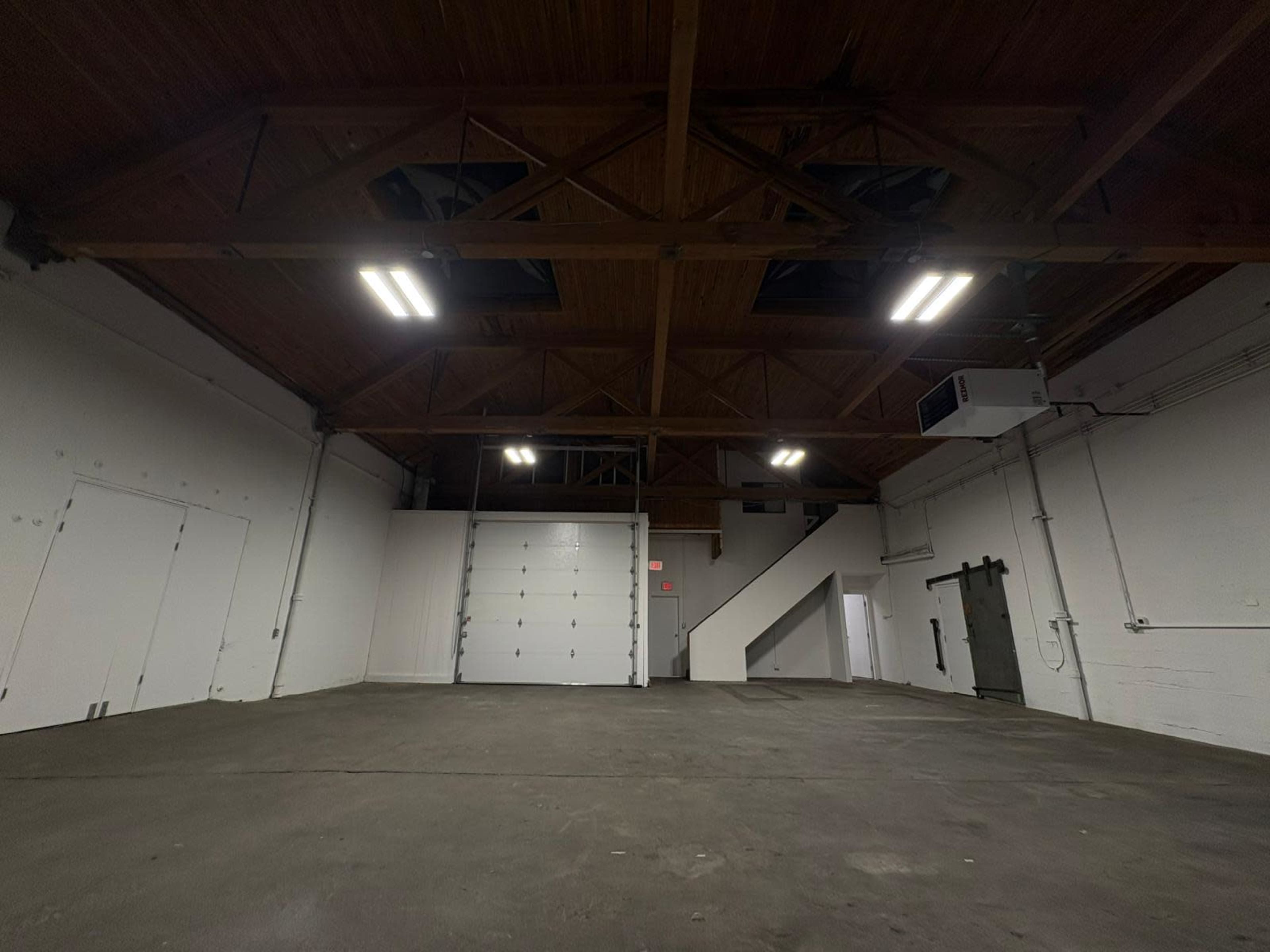 The image shows an empty industrial space with high ceilings, exposed beams, and a concrete floor, featuring a large garage door and a staircase.