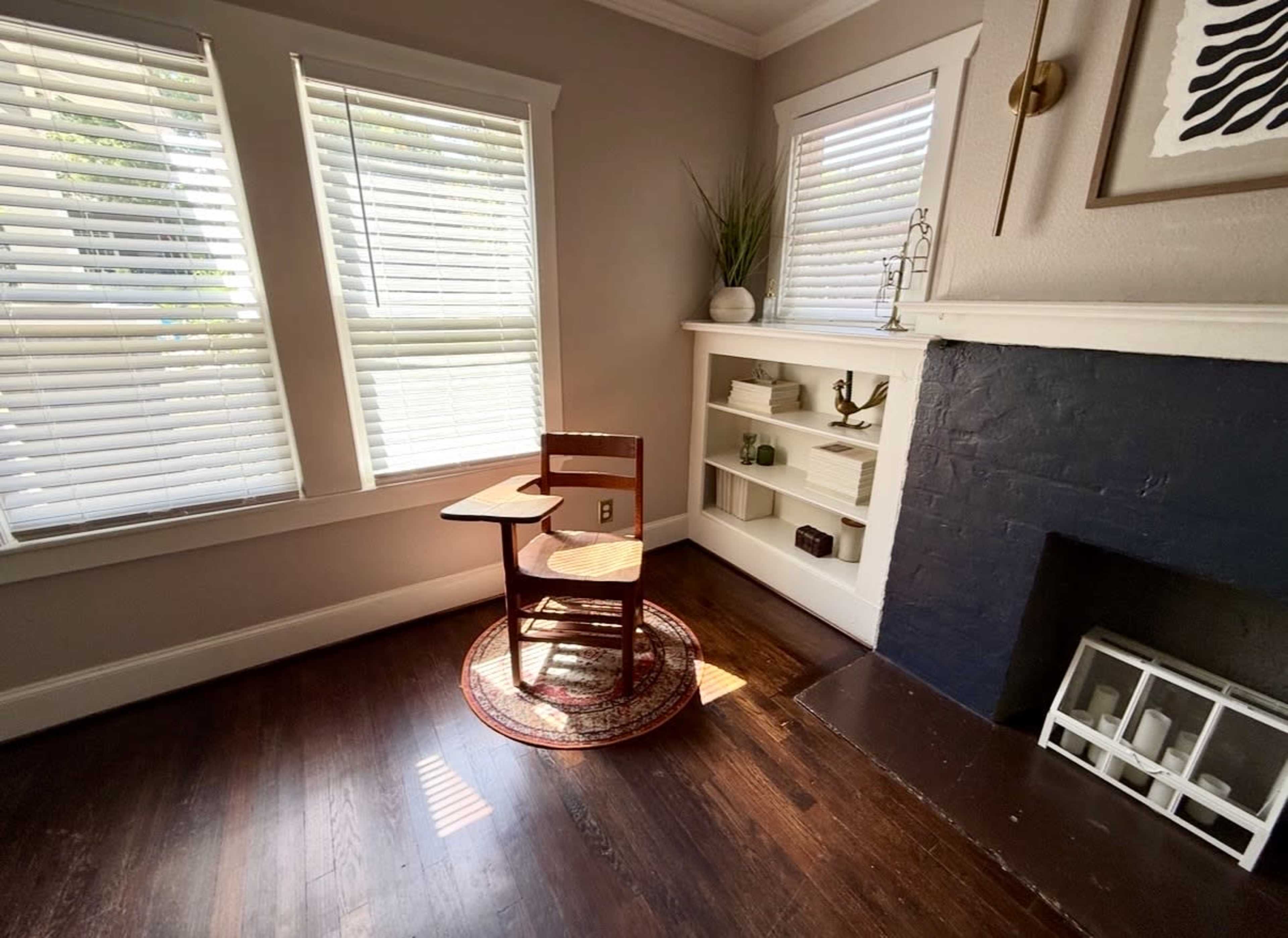 A wooden chair with a side table is positioned on a circular rug in a sunlit corner of a room featuring a fireplace and built-in shelves.