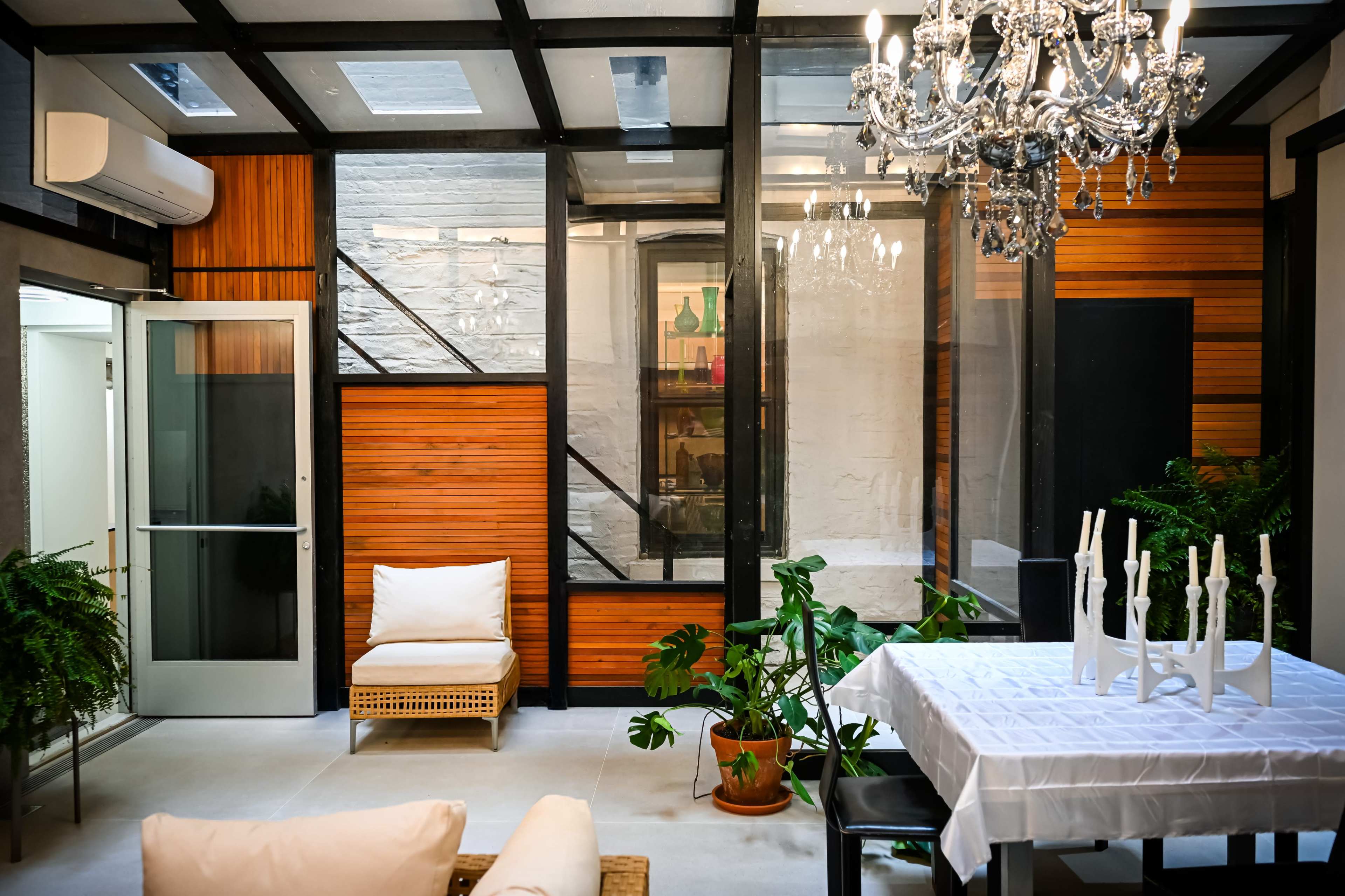 The image shows a modern living space featuring a white dining table, a chandelier, wooden accents, potted plants, and a large glass door leading outside.