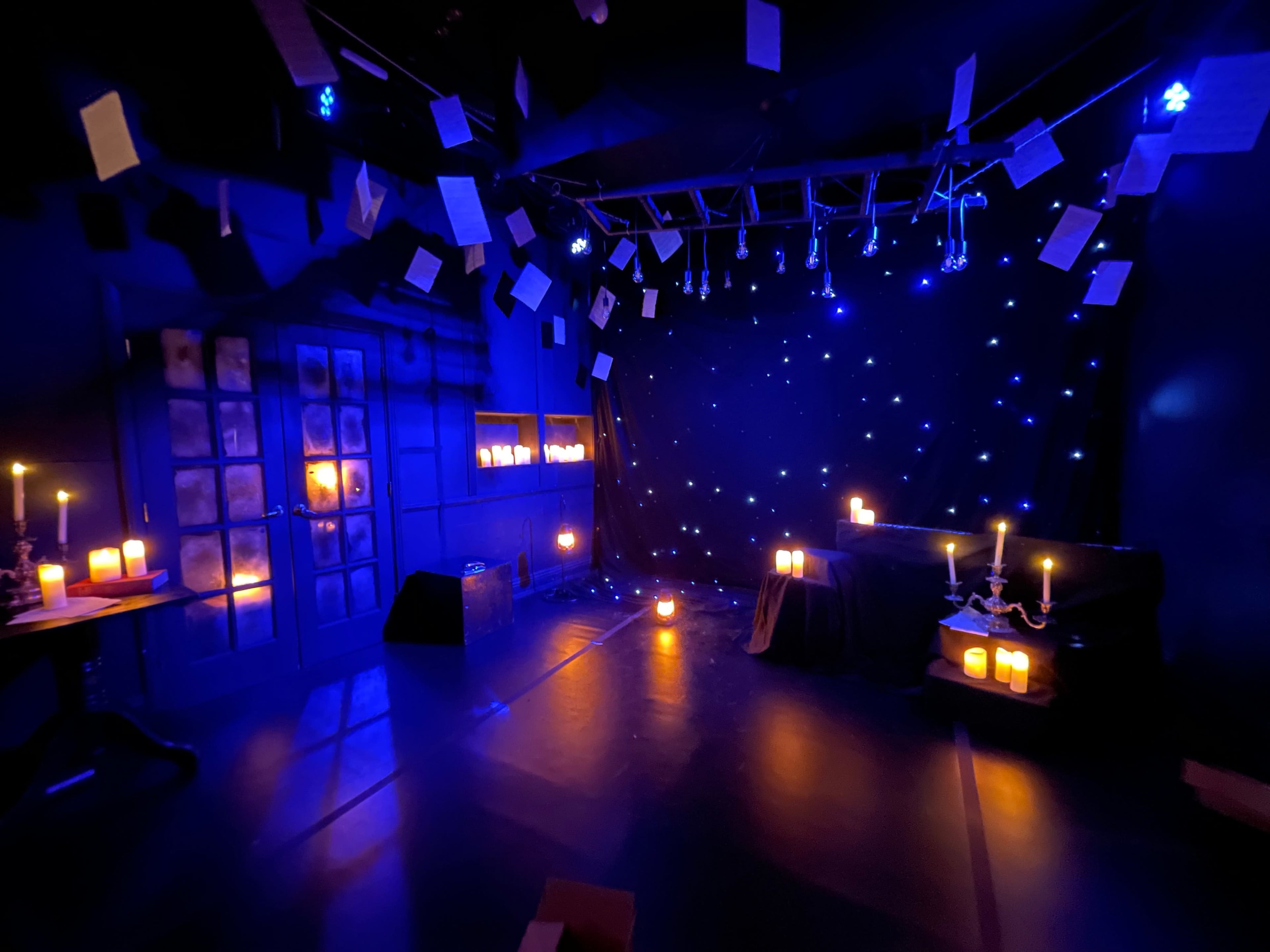 The scene depicts a dimly lit room with blue lighting, featuring candles, scattered pages hanging from the ceiling, and a table set against a starry backdrop.