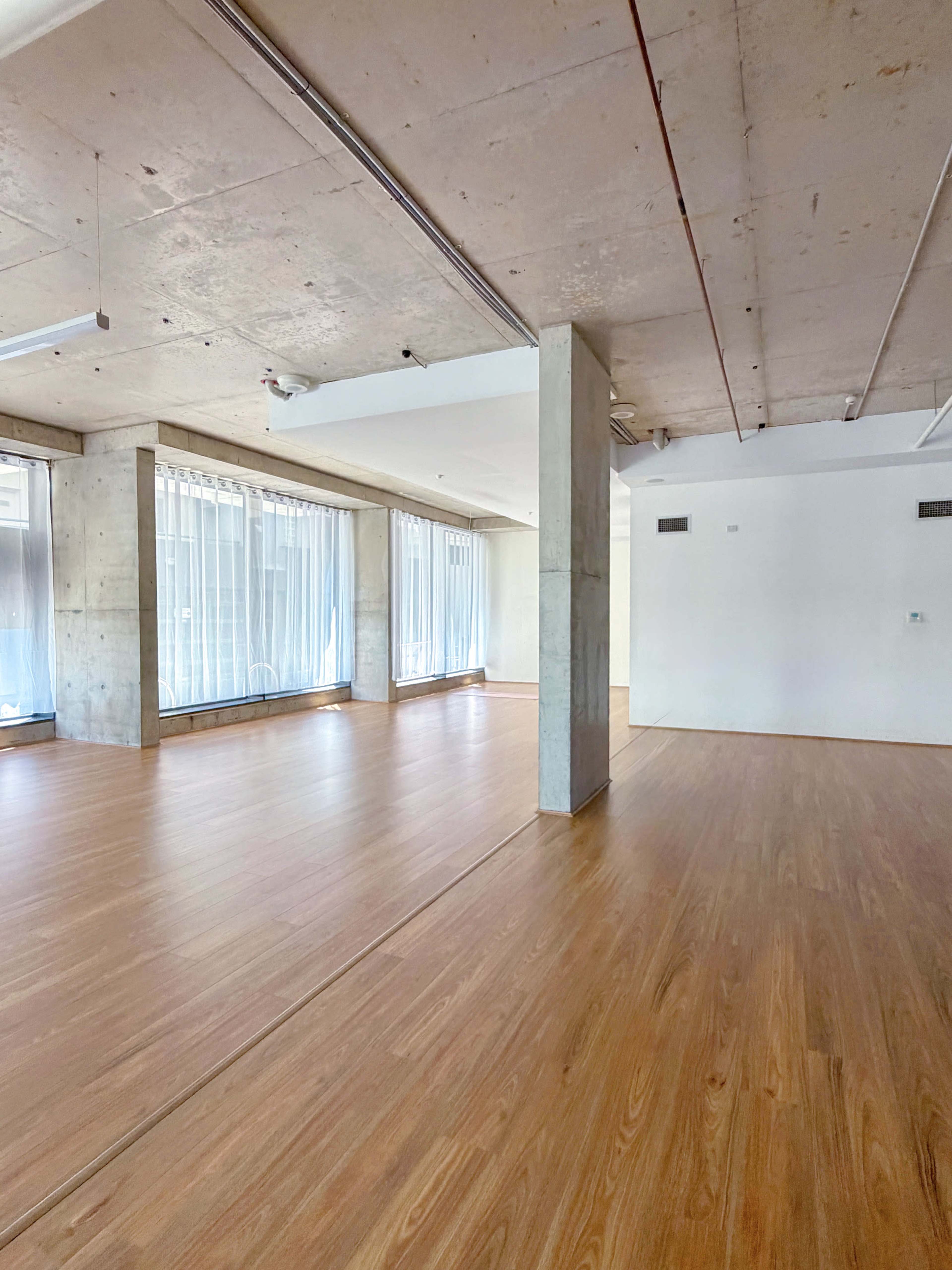 The image shows a spacious, empty room with large windows, concrete pillars, and polished wooden flooring.
