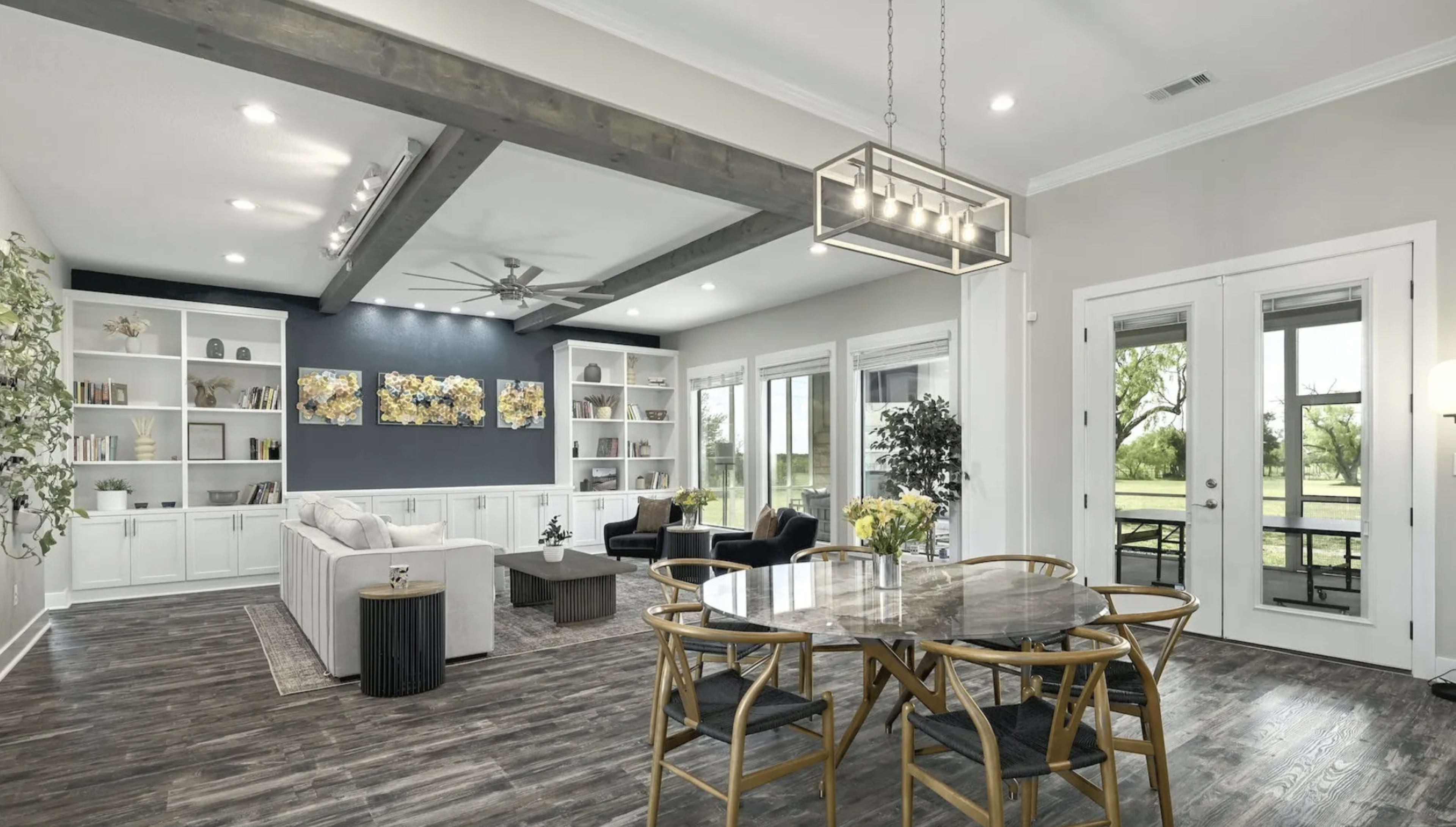 A modern living room and dining area with a light-colored sectional sofa, a circular dining table, and large windows that provide views of the outdoors.