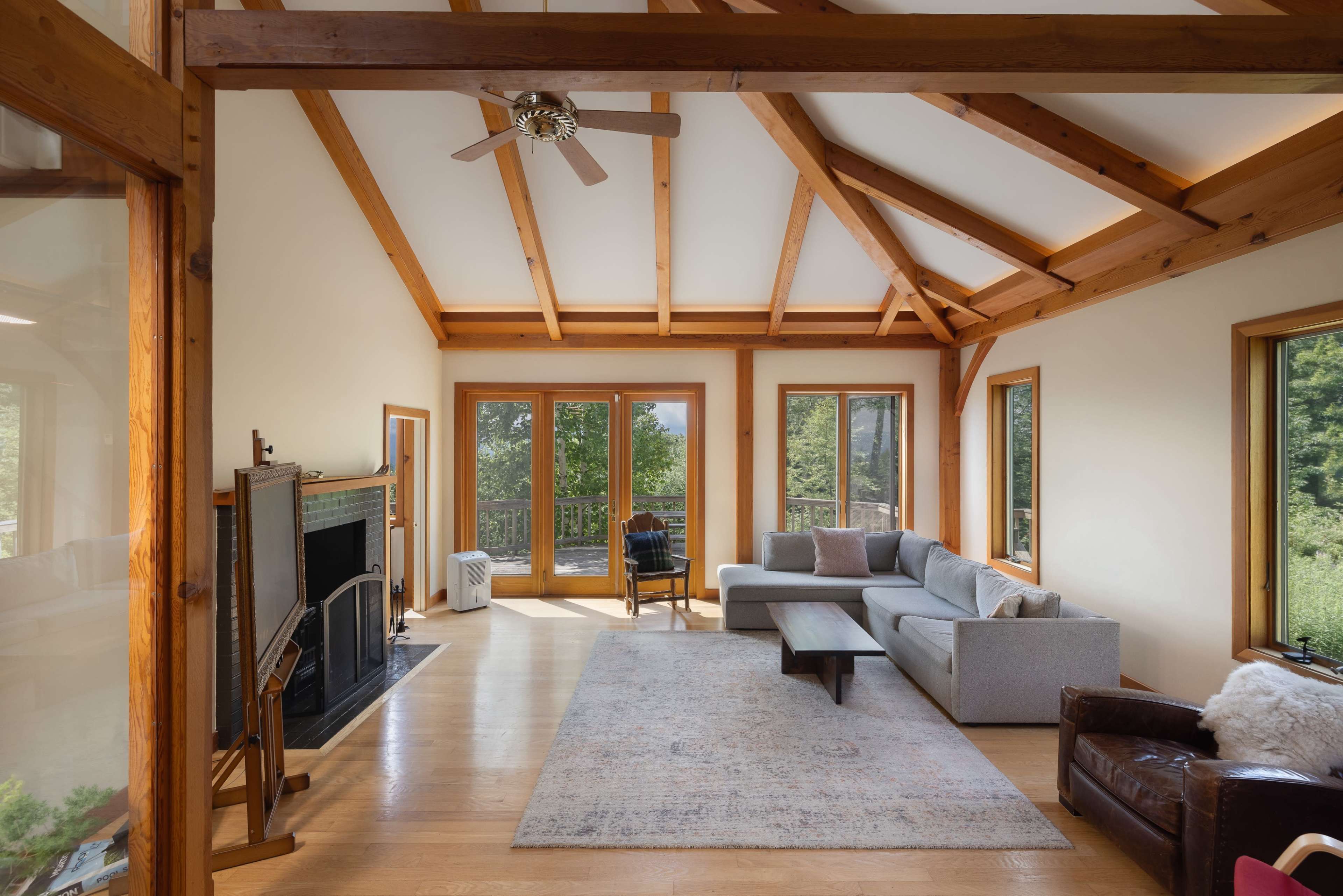 The image shows a living room featuring wooden beams, large windows, a gray sectional sofa, a wooden coffee table, and a fireplace.