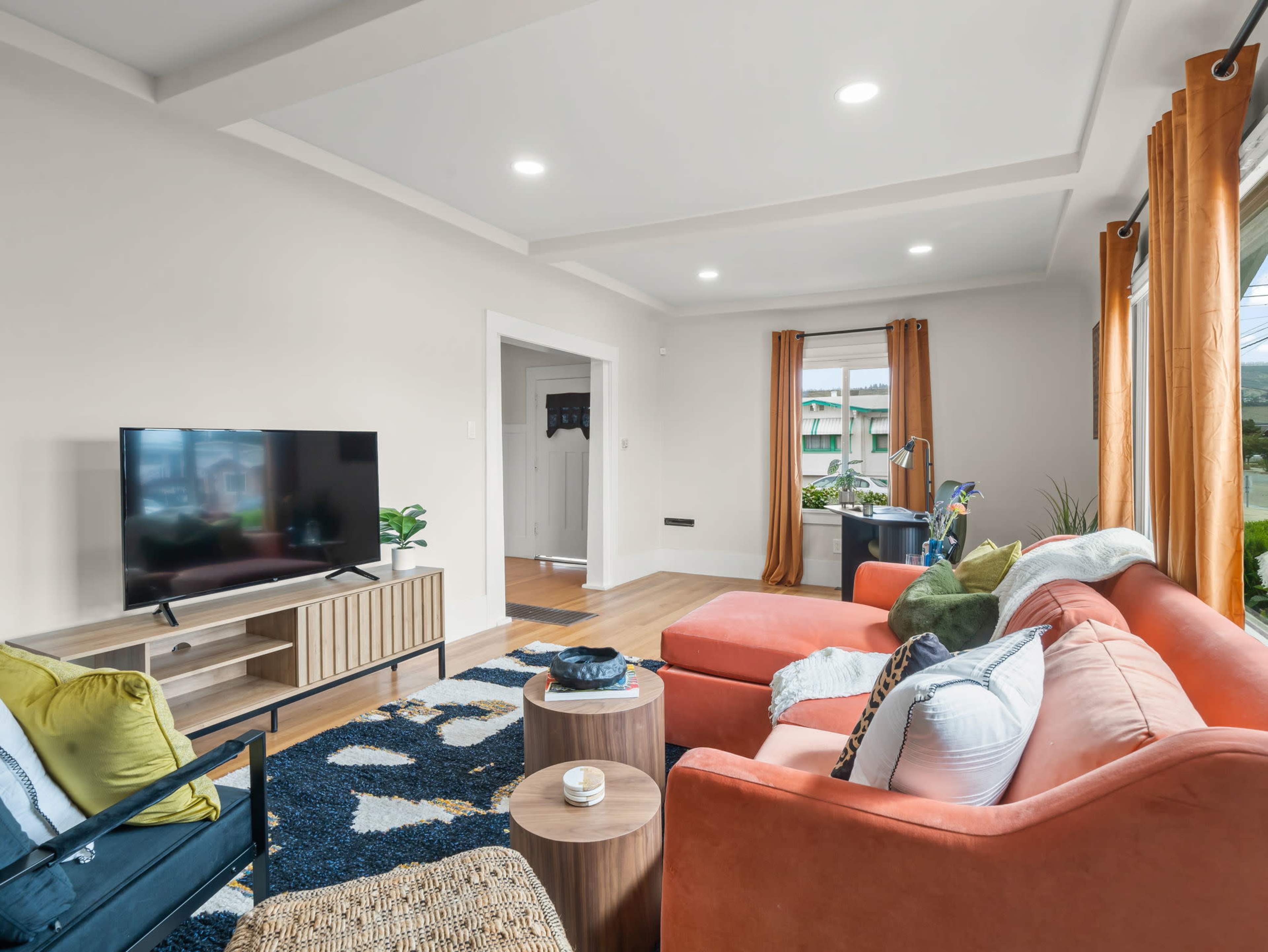 The living room features an orange sofa, a television on a wooden stand, and large windows with orange curtains, creating a bright and inviting space.