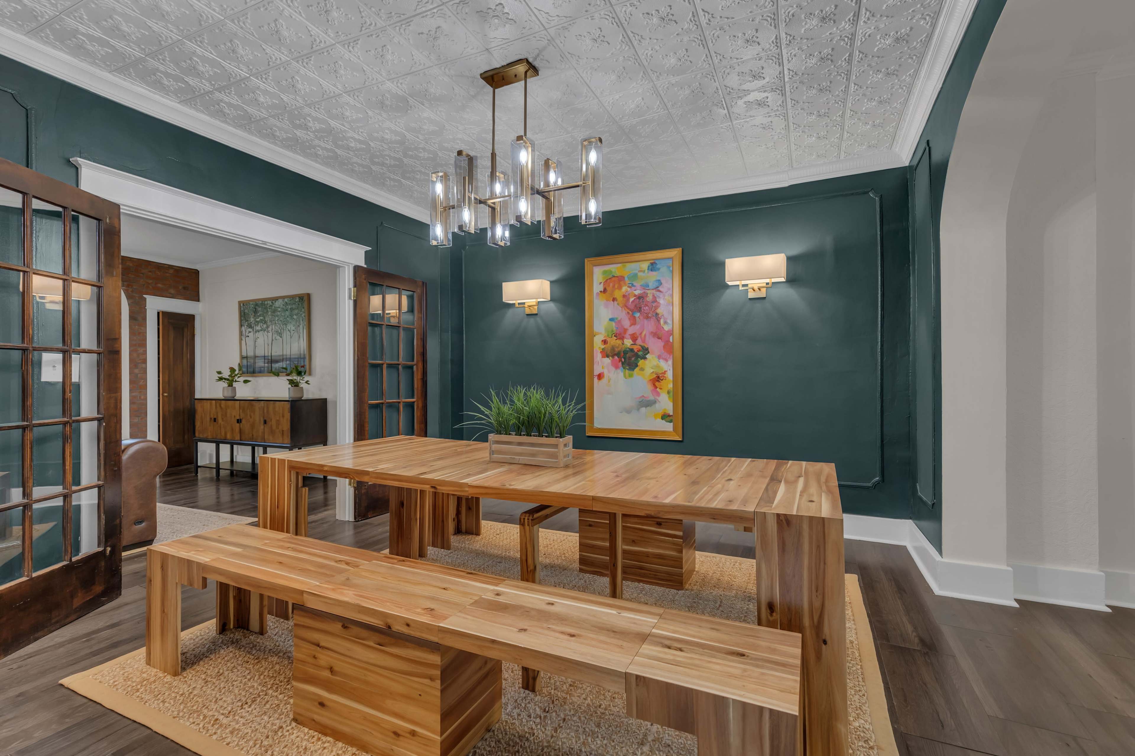 A spacious dining room features a large wooden table with matching benches, green walls, and a modern chandelier.