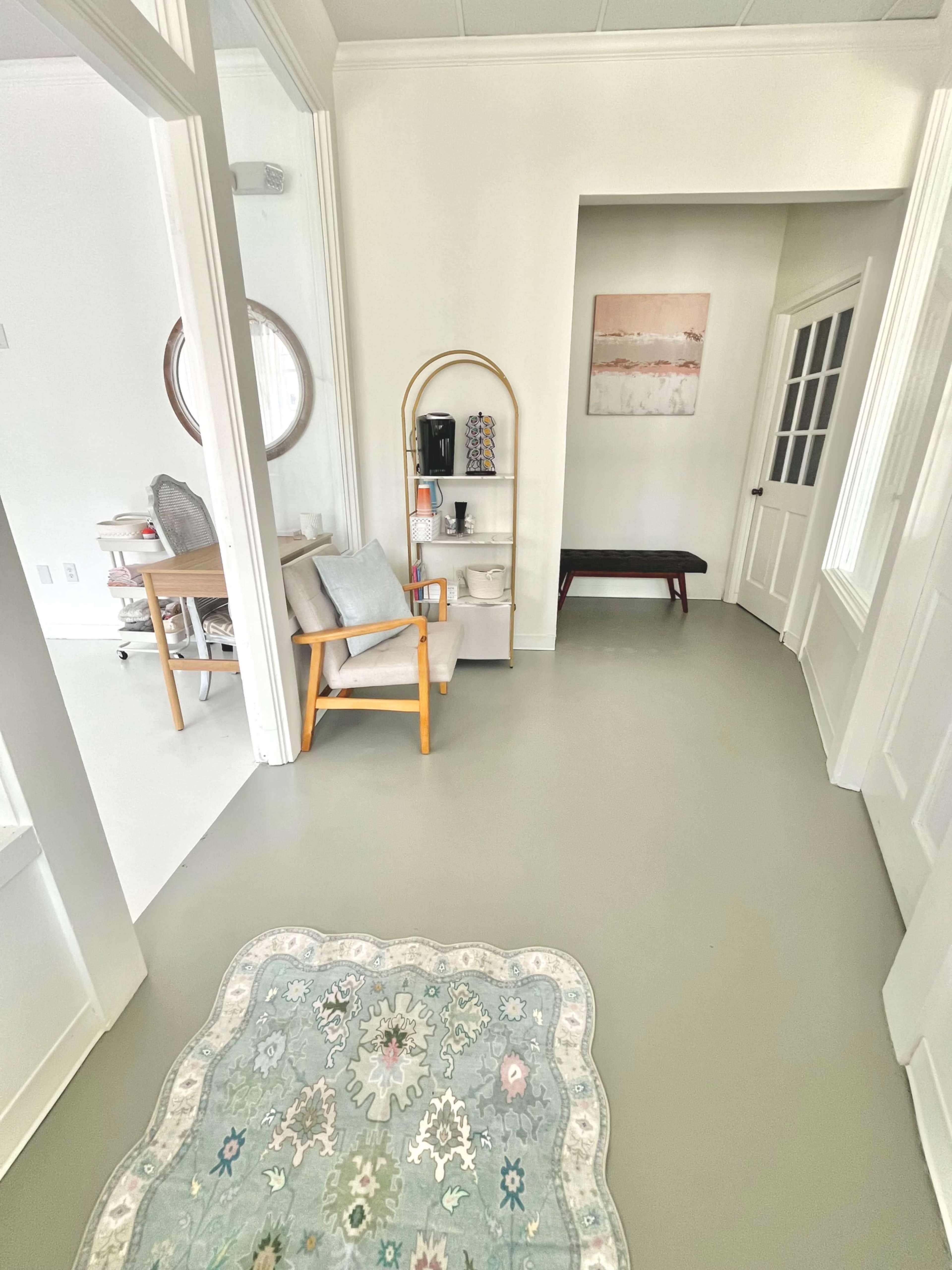 The image shows a bright hallway featuring a patterned rug, a wooden bench, a shelving unit with decorative items, and a chair beside a door leading to another room.