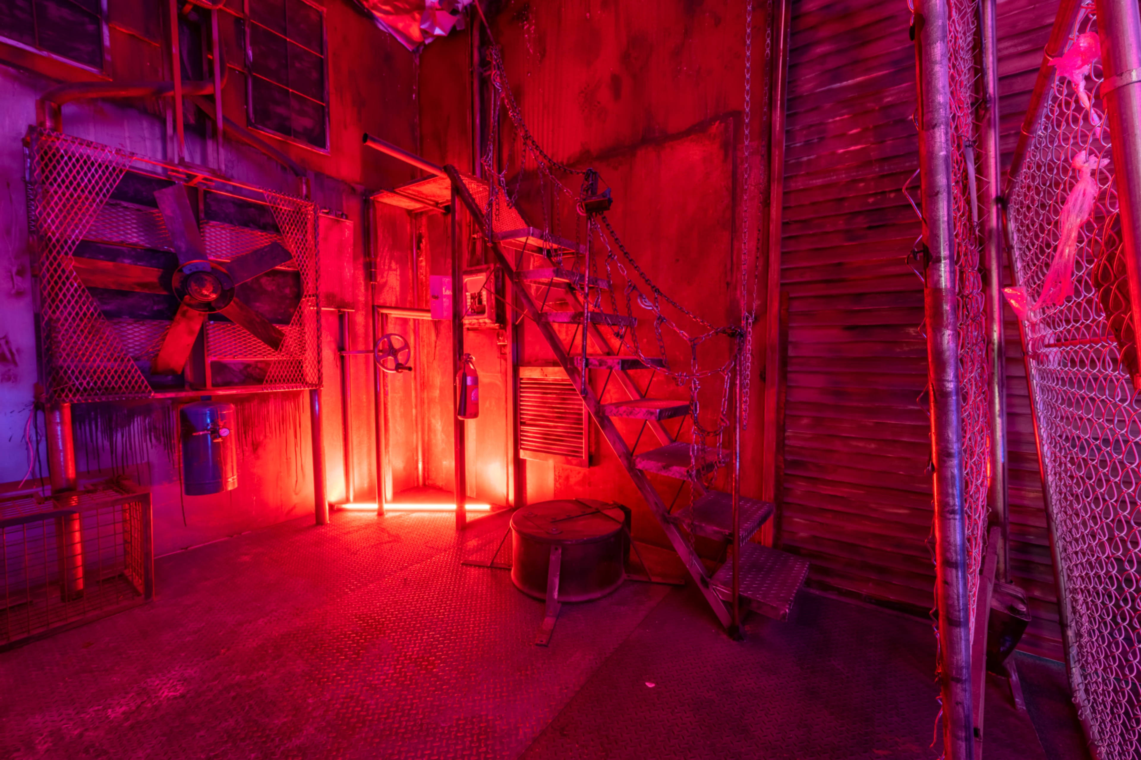 A dimly lit industrial space features a metallic staircase, chains, and large fans against walls painted in shades of red.