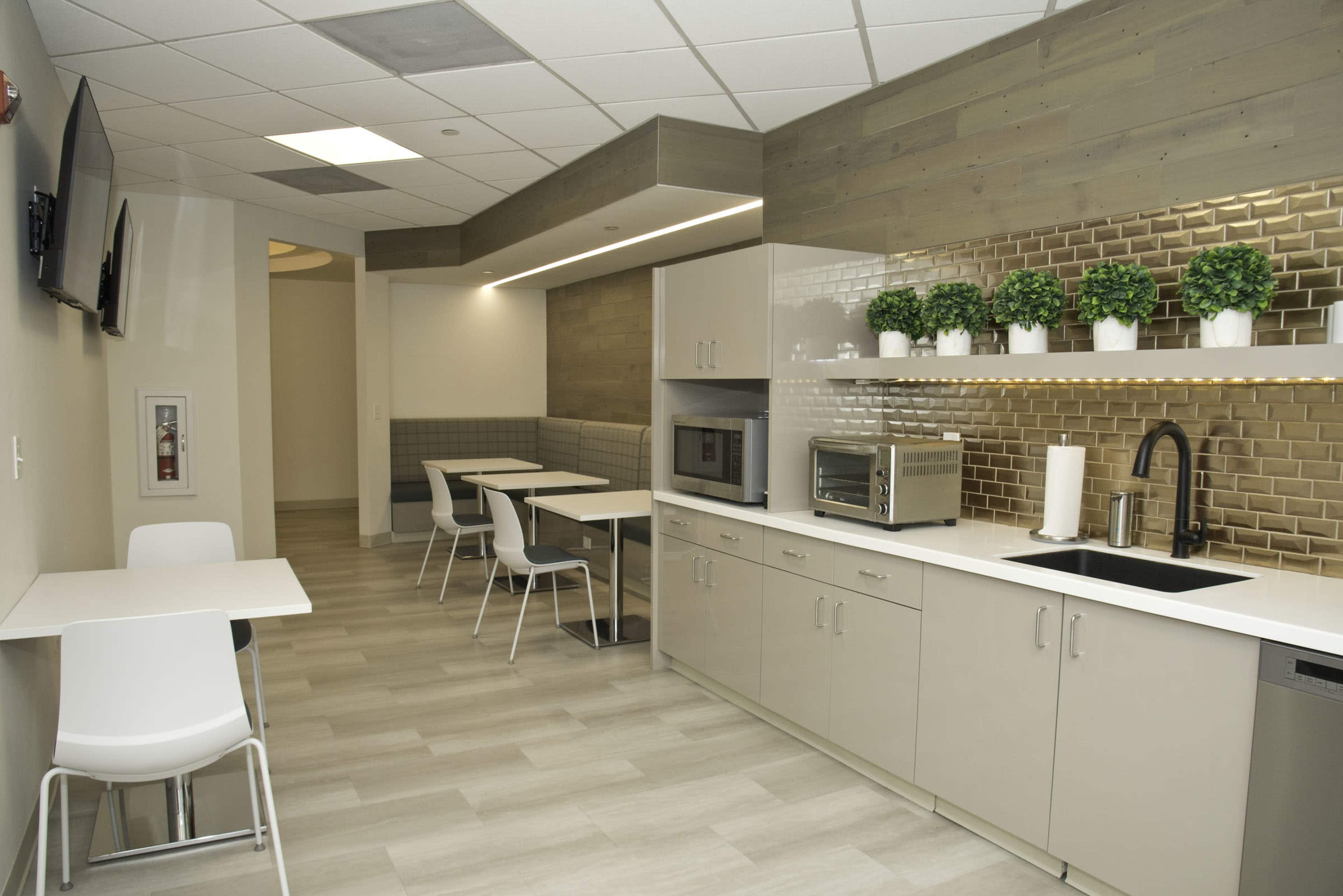 A modern kitchen area features white cabinetry, a microwave, a sink, and tables surrounded by chairs, with potted plants on the countertop.