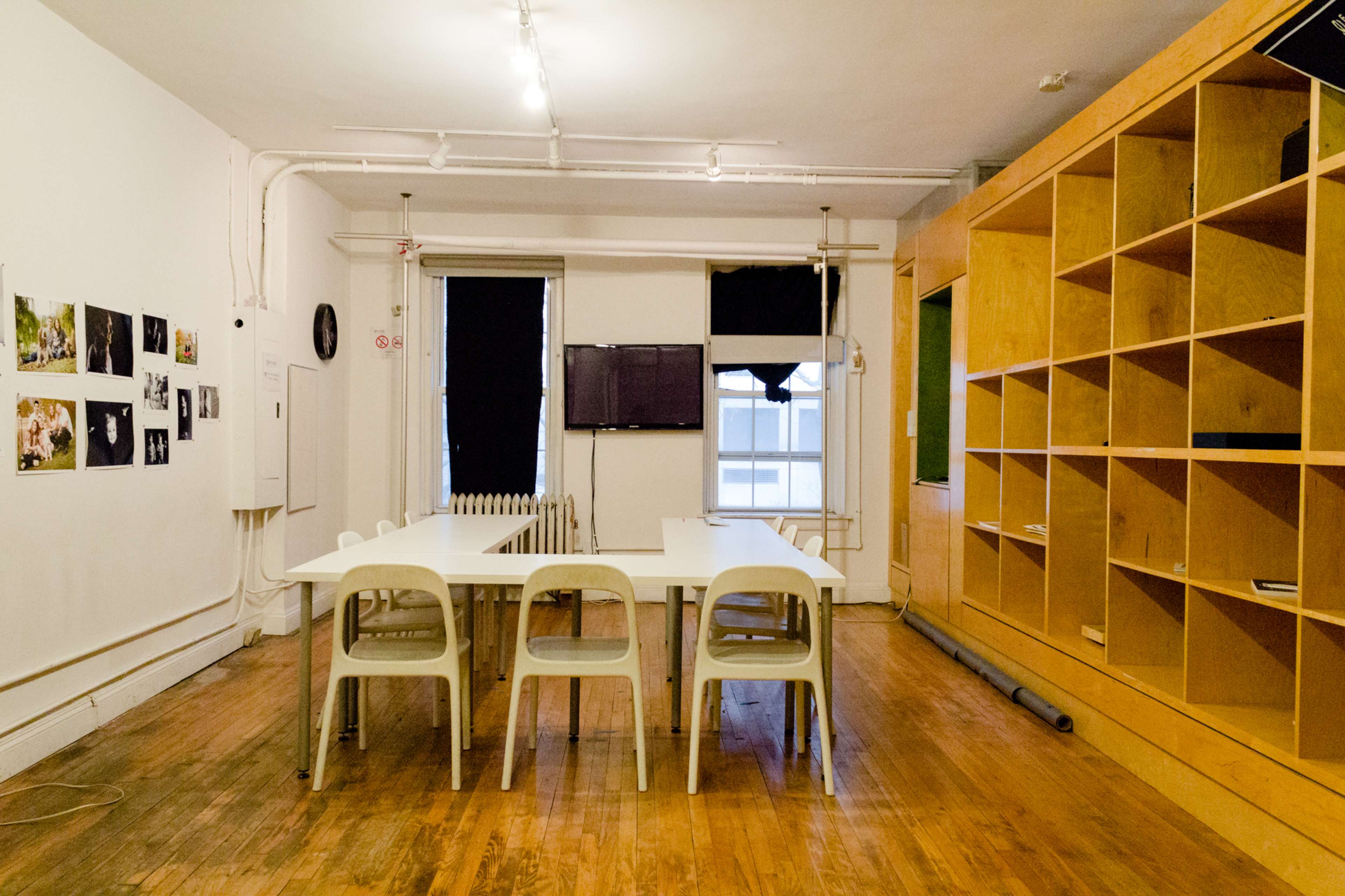 A spacious room features a long table surrounded by plastic chairs, with wooden shelves and a television mounted on the wall.