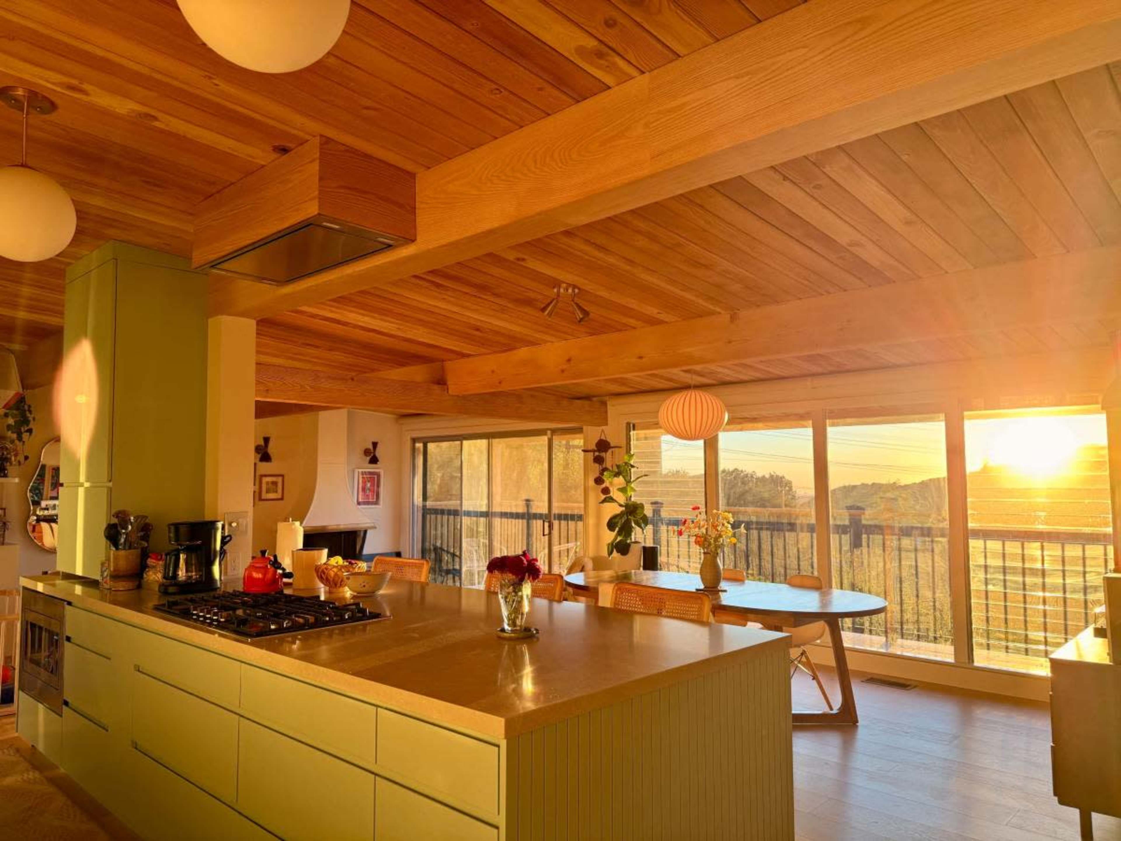 A modern kitchen with a green island and wooden beams features large windows that overlook a sunset.