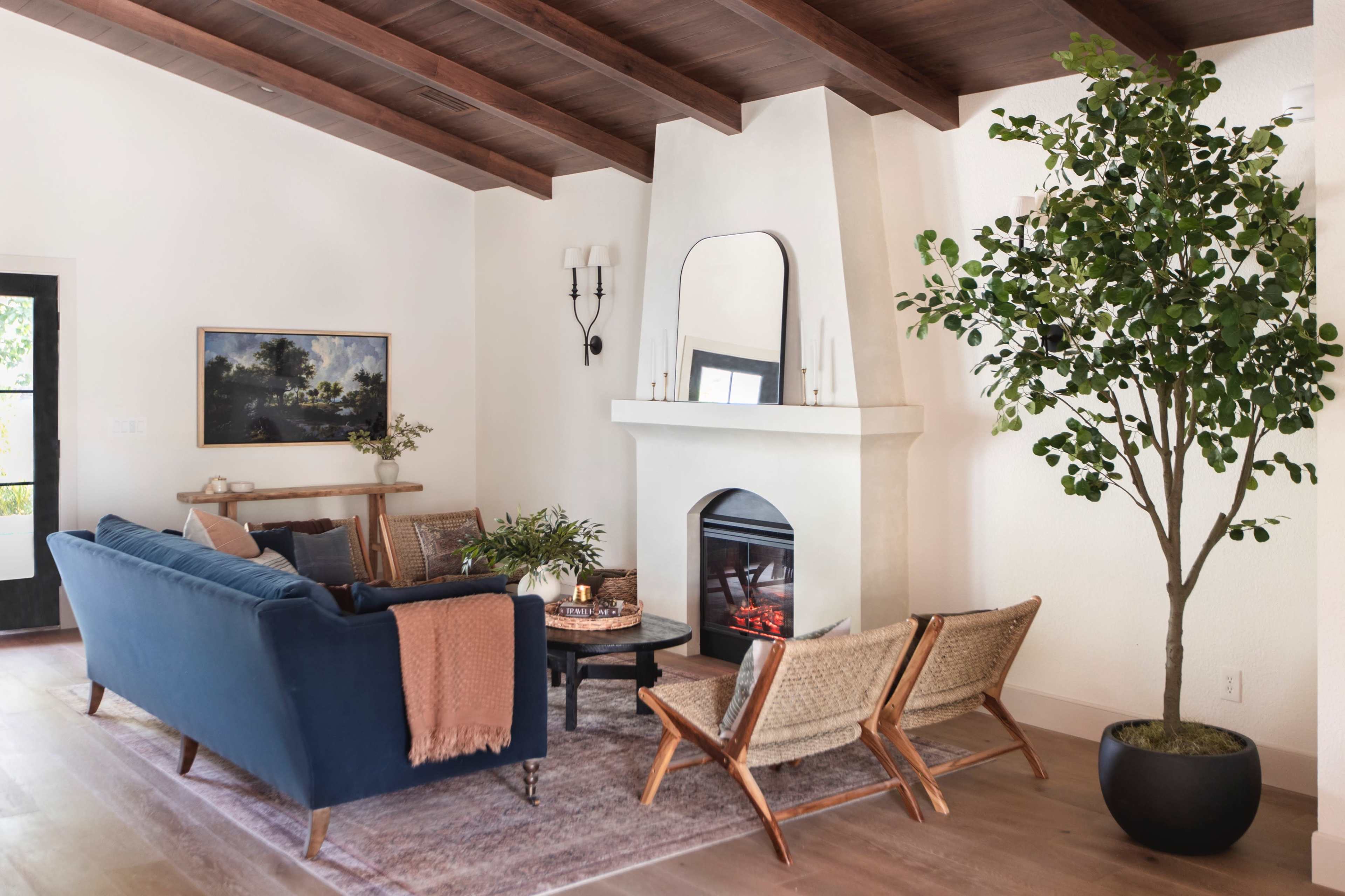 A modern living room featuring a blue sofa, two woven chairs, a fireplace, and a large potted plant beside a beige wall with wooden beams.