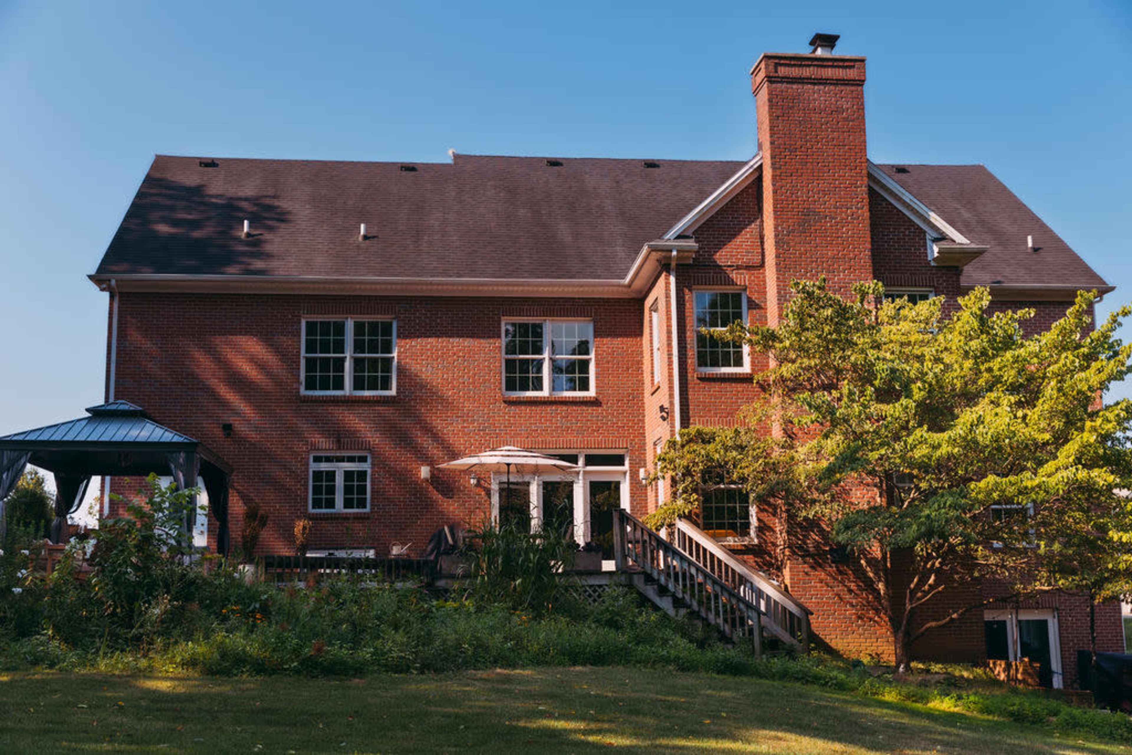 A large brick house with a chimney and multiple windows is surrounded by a grassy yard and a tree.