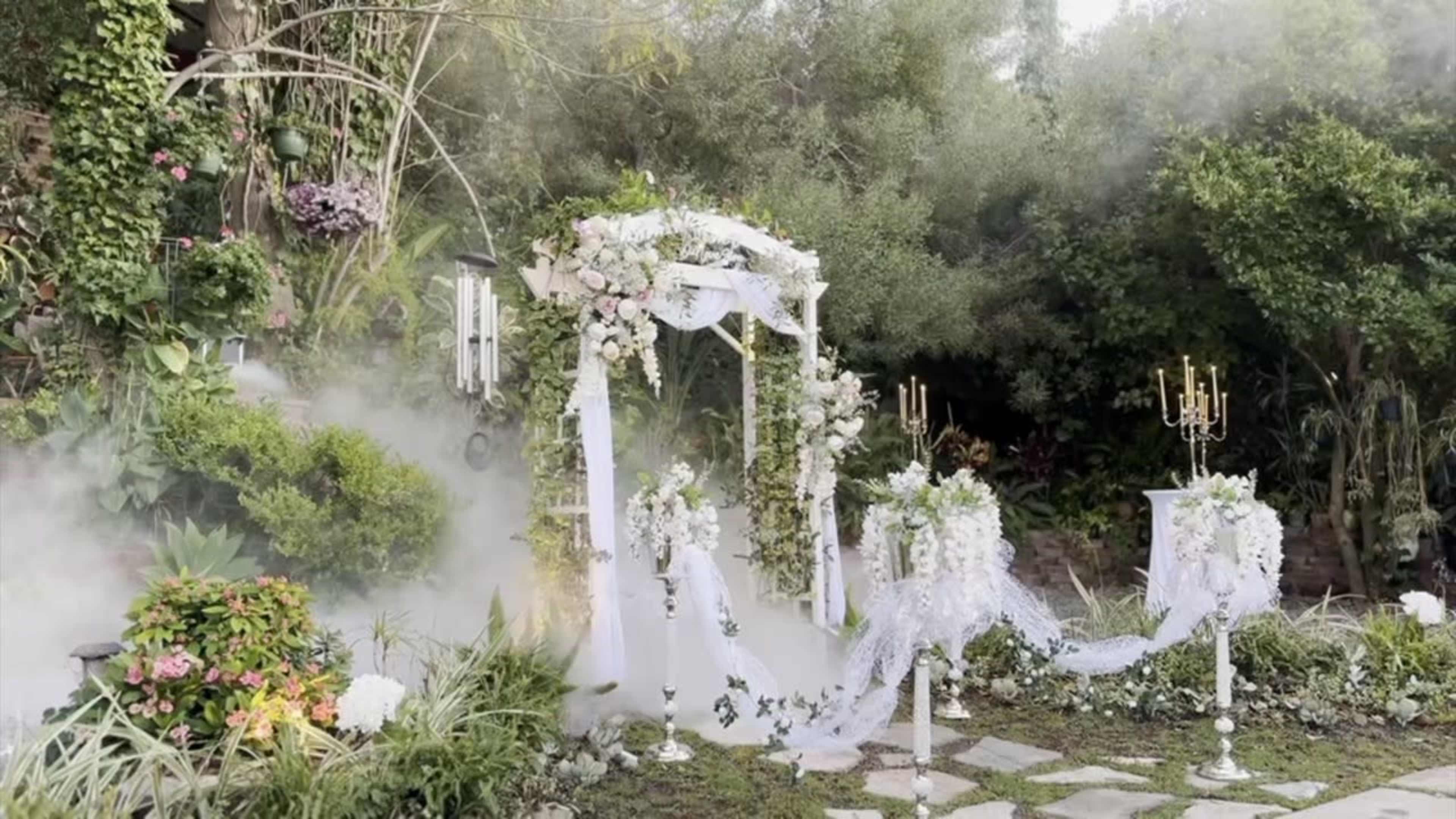A decorative wedding altar is set up in a lush garden, surrounded by greenery and adorned with flowers, with mist gently enveloping the scene.