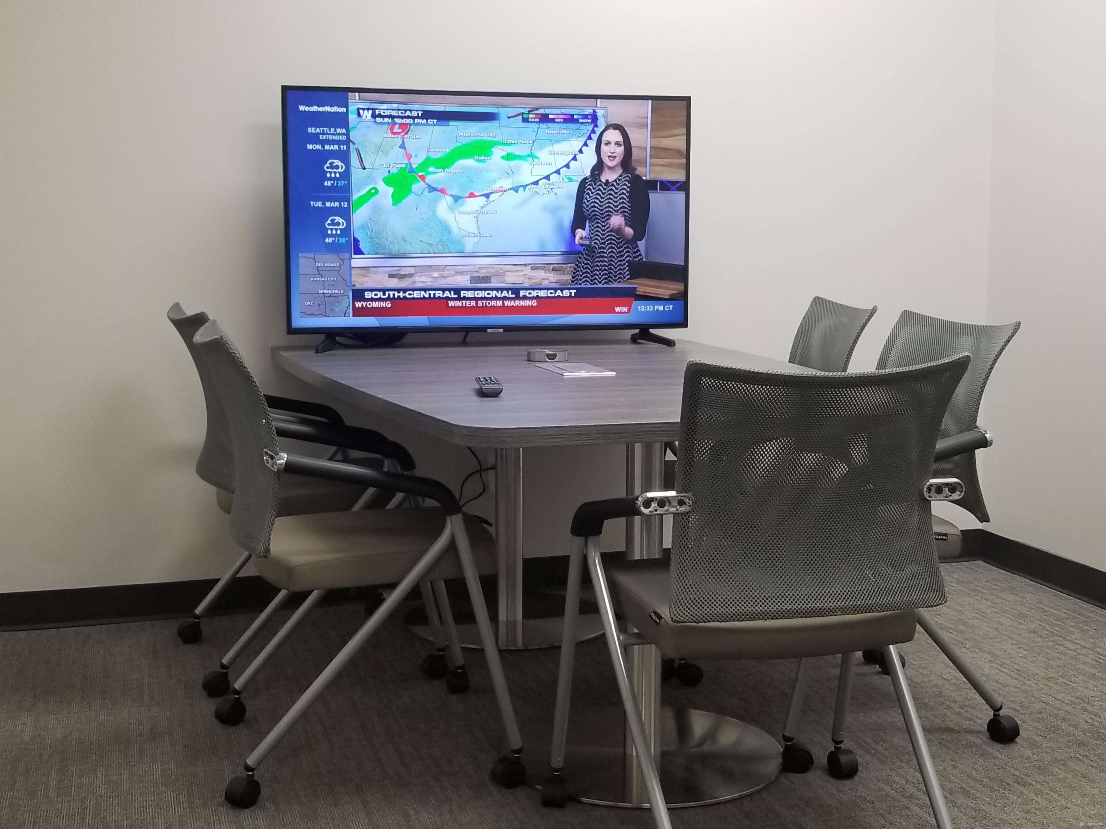 A conference room features a large television displaying a weather forecast, with several chairs arranged around a table.