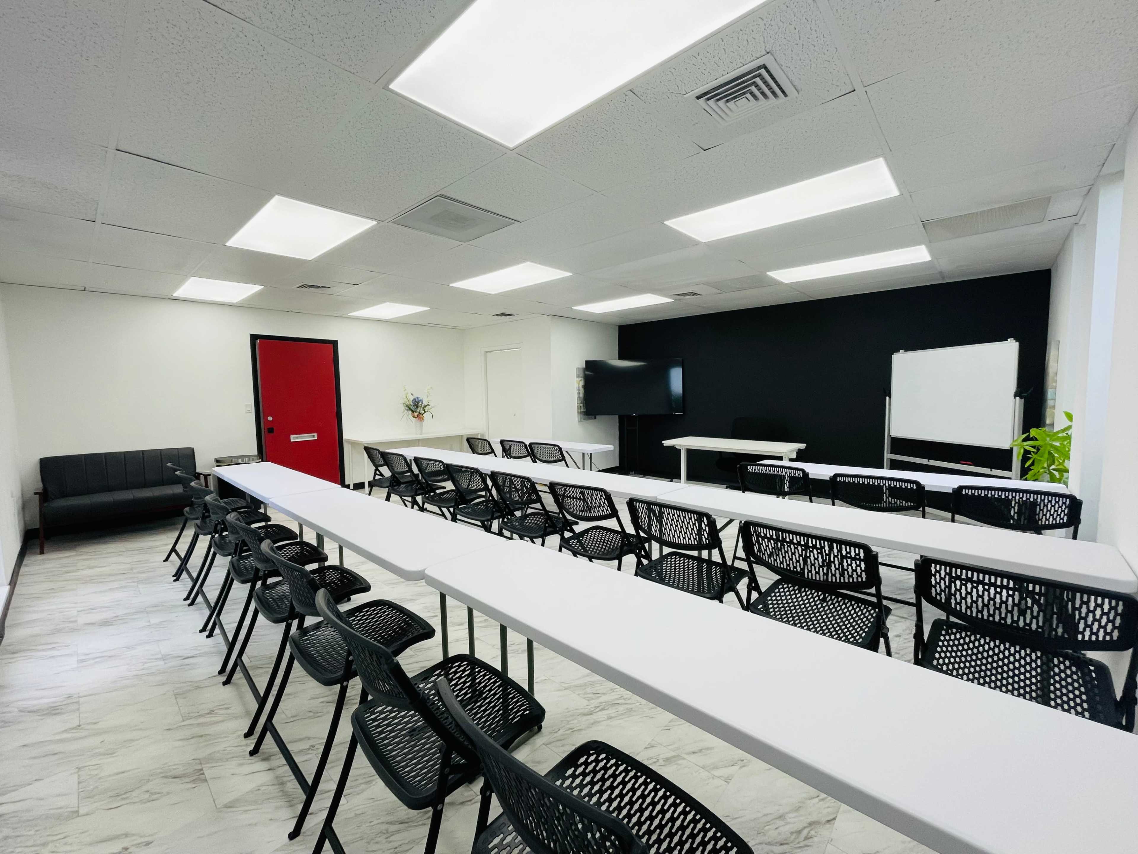 The image shows a spacious conference room with rows of black chairs facing long white tables, a black wall with a whiteboard, and a red door.