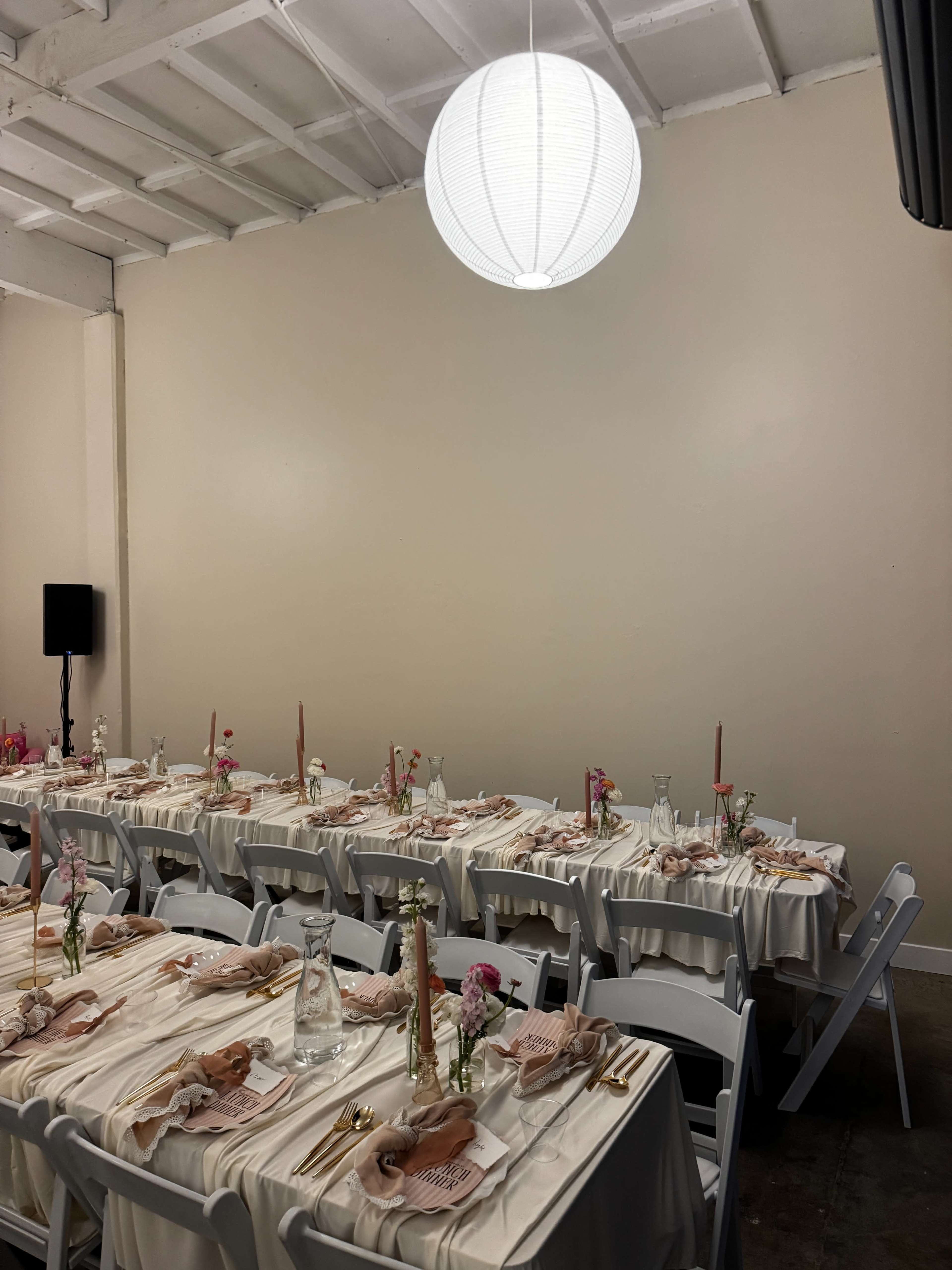 A dining area is set up with tables covered in white linens, adorned with pink napkins, flowers, and gold utensils, under a round hanging light fixture.