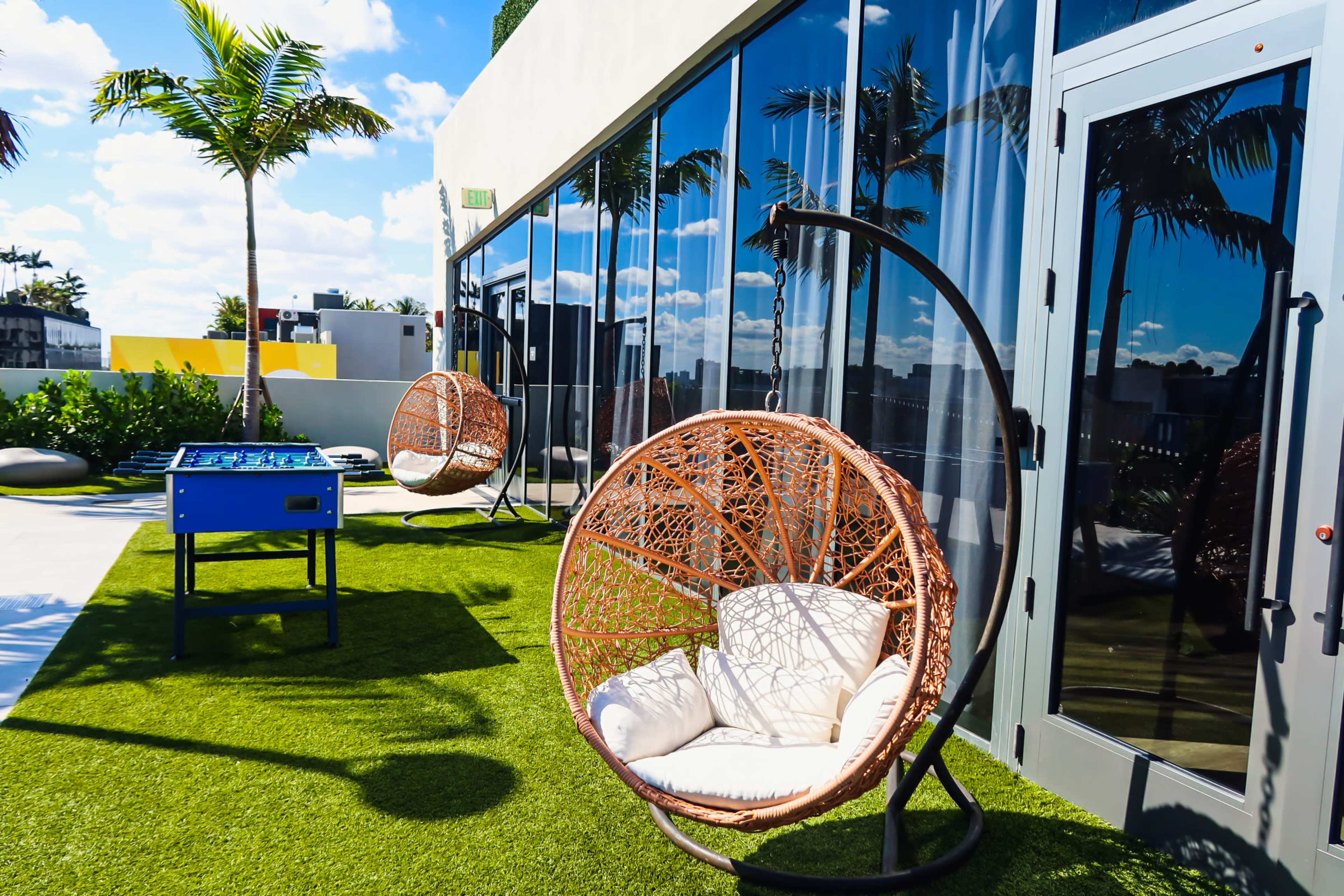 The image shows a modern rooftop terrace with two round hanging chairs and a foosball table on artificial grass under a bright sky.