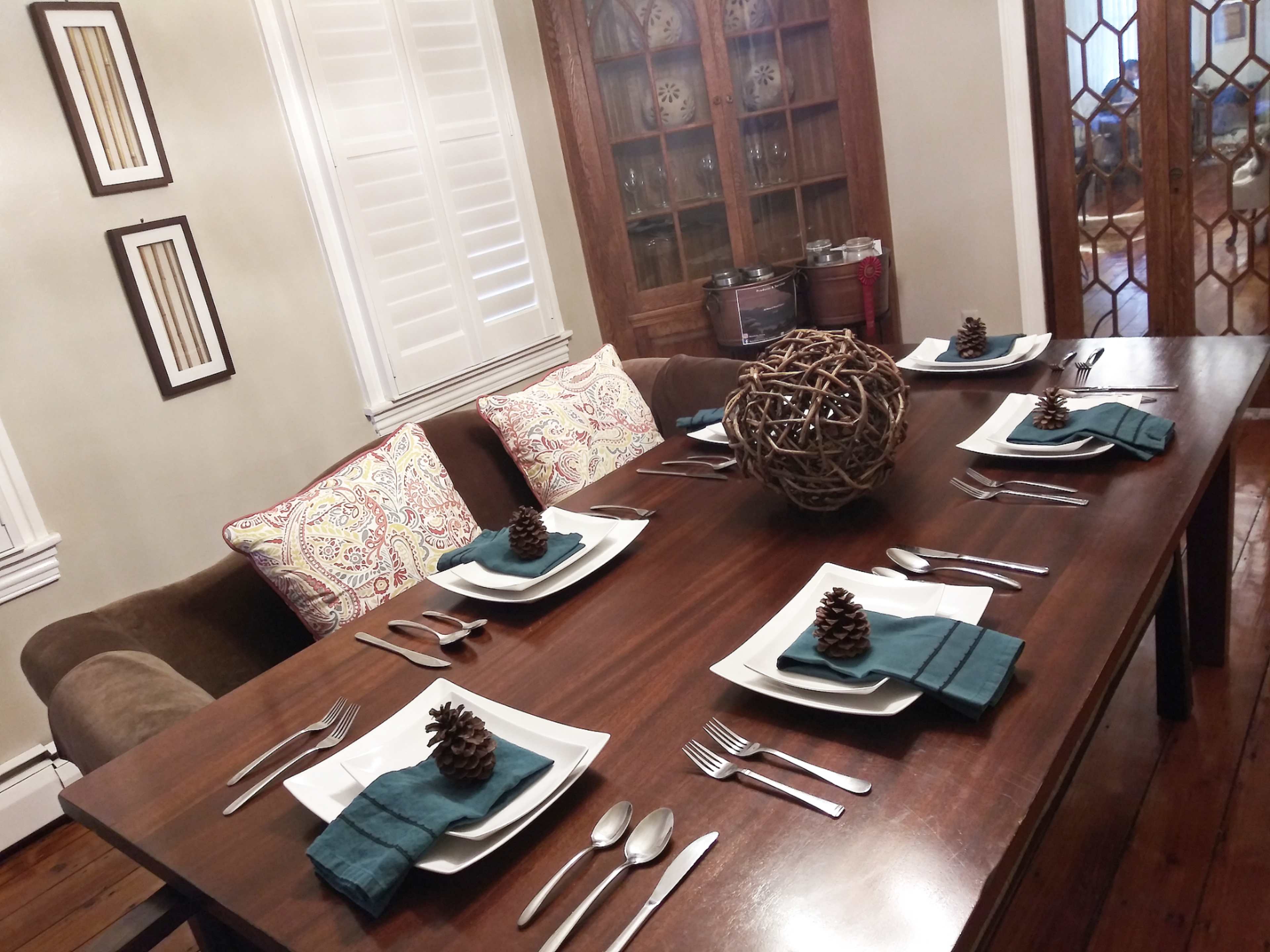 A dining table is set with plates, utensils, and decorative pinecones, surrounded by cushioned chairs and a cabinet with glassware in the background.