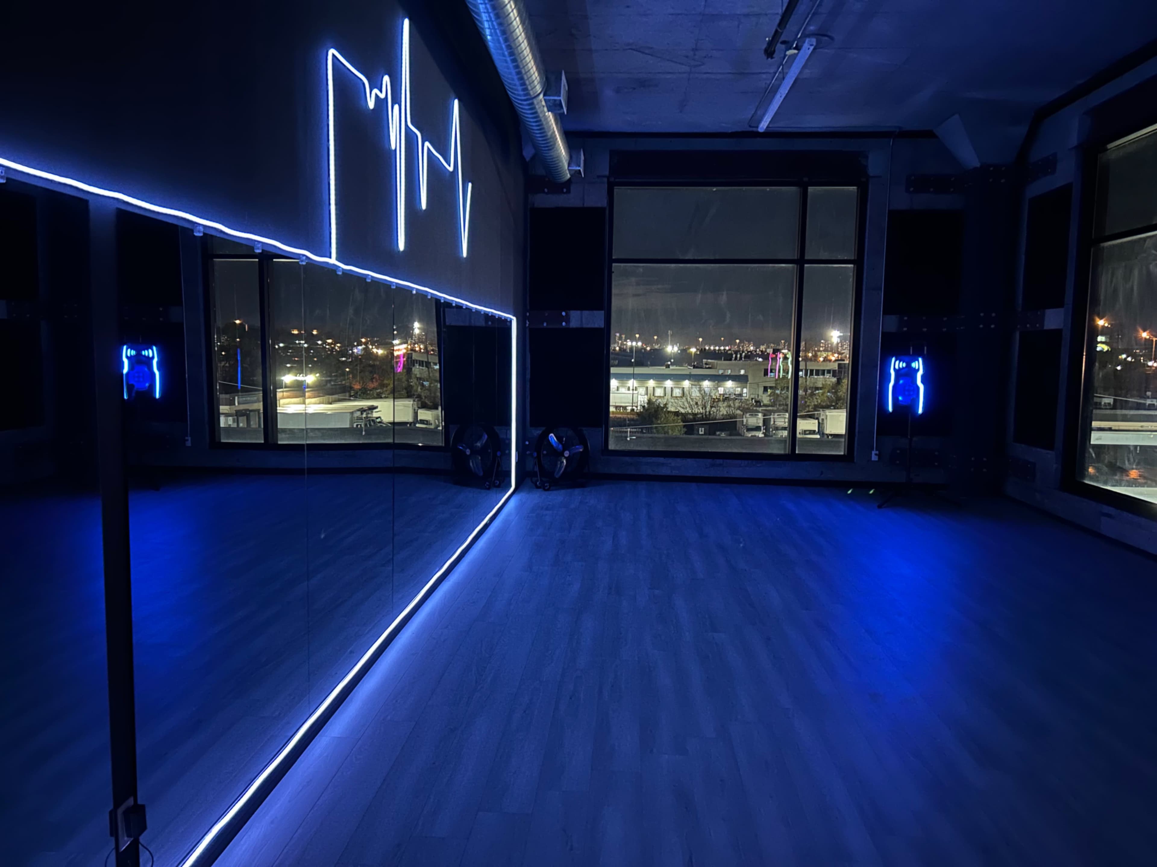 The image shows a dance studio illuminated by blue LED lights, featuring a large mirror and a city skyline visible through the window.
