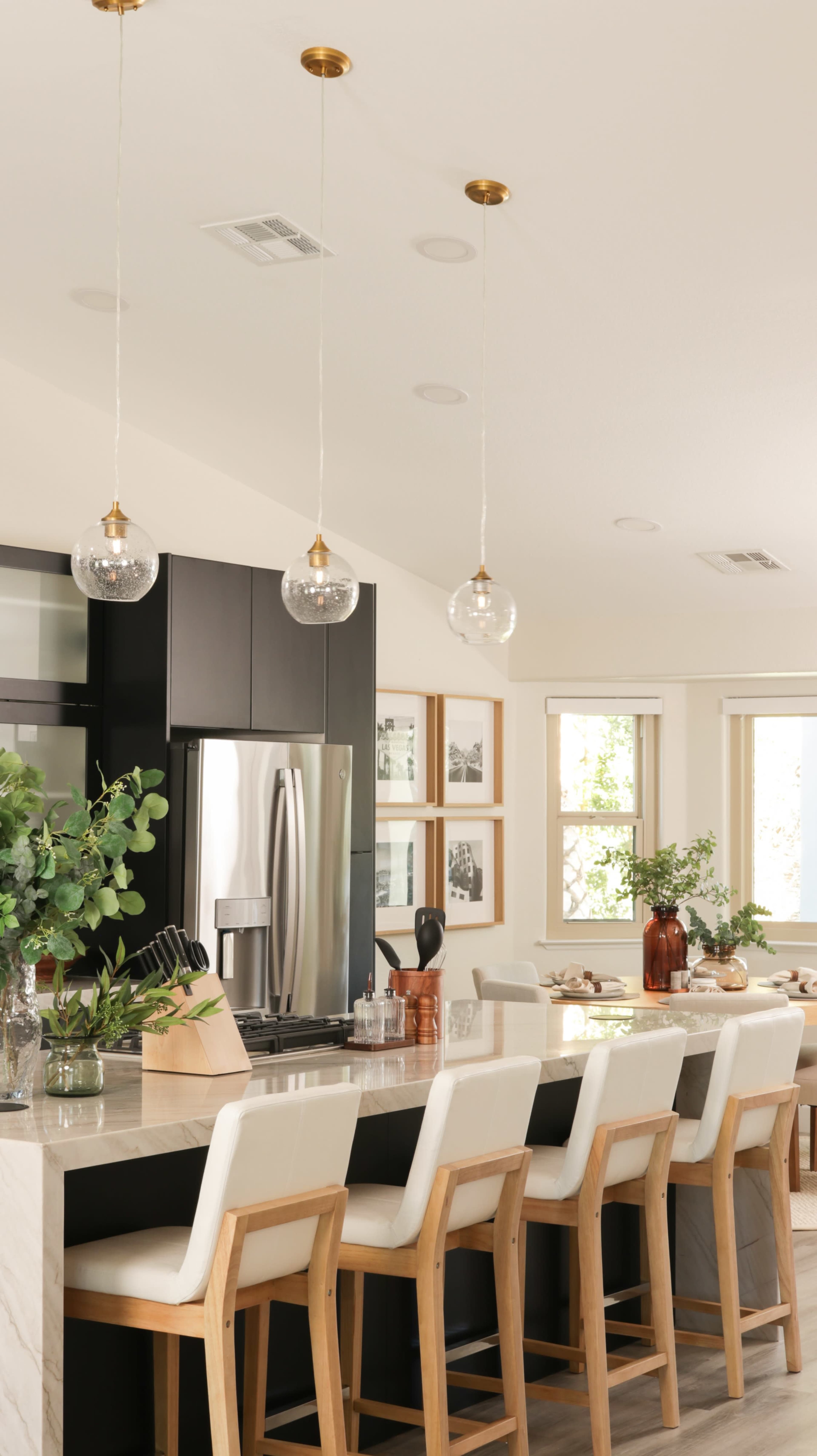 A modern kitchen features a large island with bar stools, sleek black cabinetry, a stainless steel refrigerator, and pendant lighting above.
