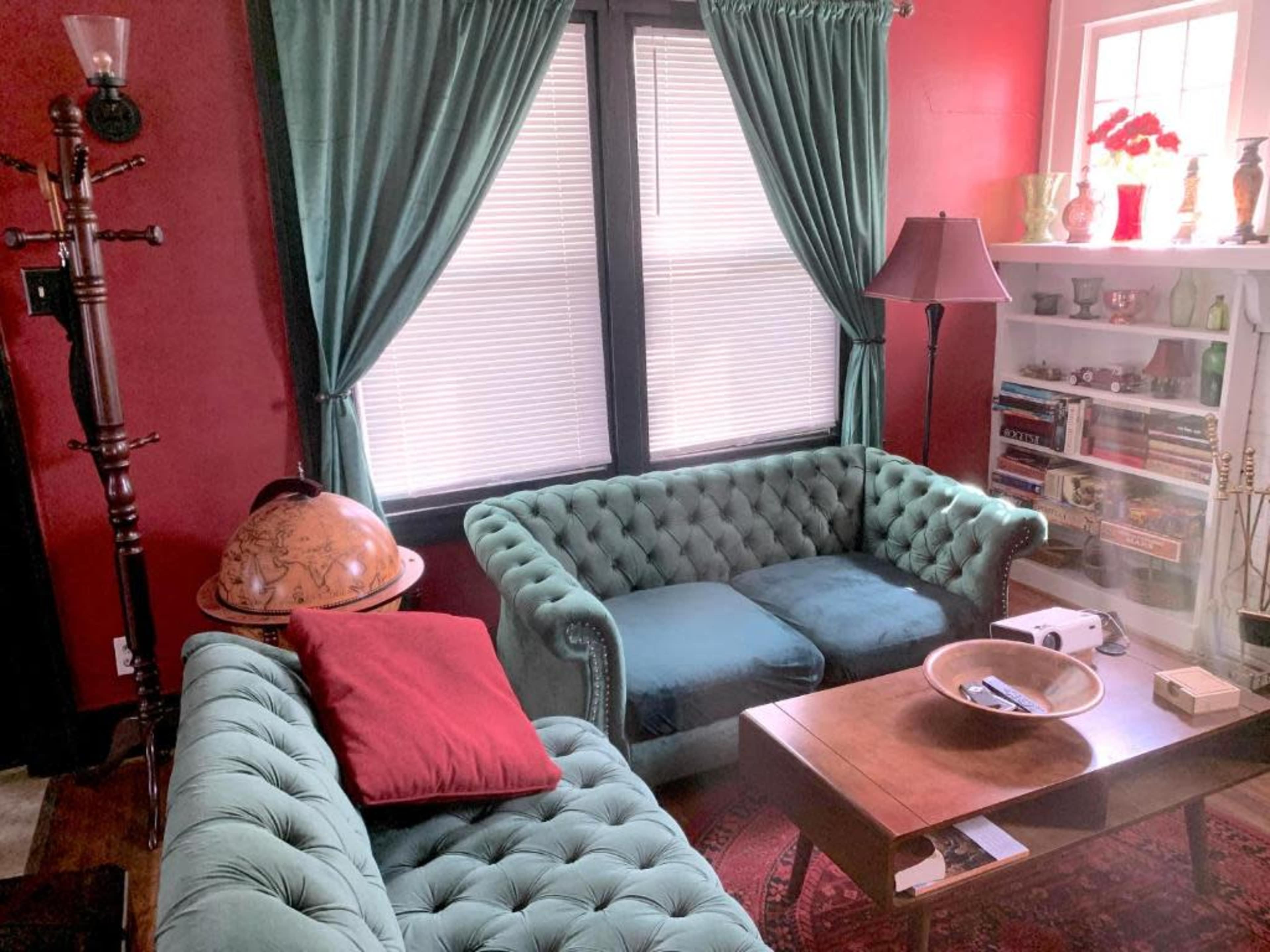 The image shows a cozy living room featuring two tufted couches, a wooden coffee table, and a globe on the side, with red walls and large windows dressed with green curtains.