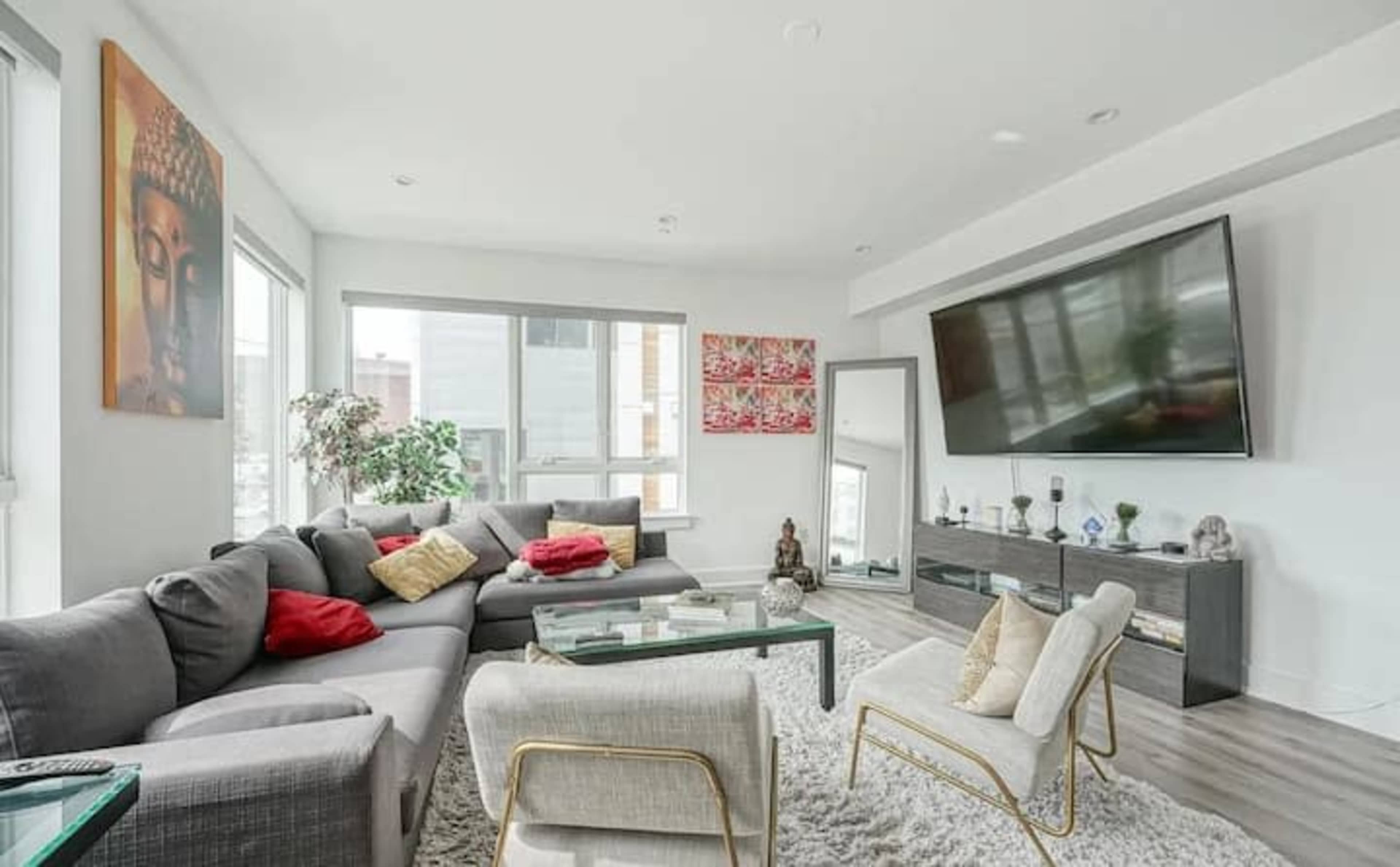 A modern living room features a gray sectional sofa, a large television mounted on the wall, and decorative artwork, with light streaming through multiple windows.
