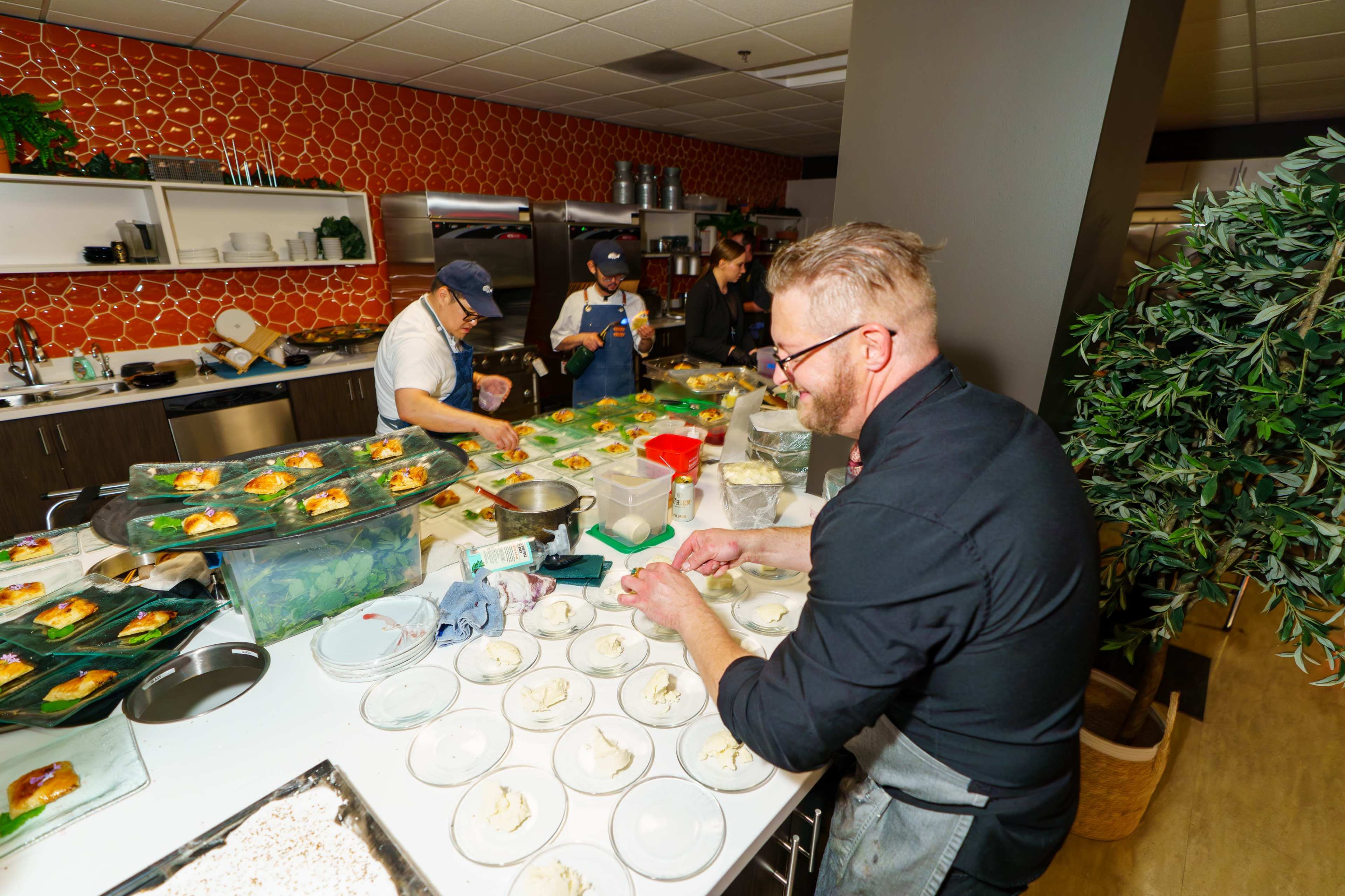 Chefs prepare plates of food in a commercial kitchen with various ingredients and dishes laid out on the counter.