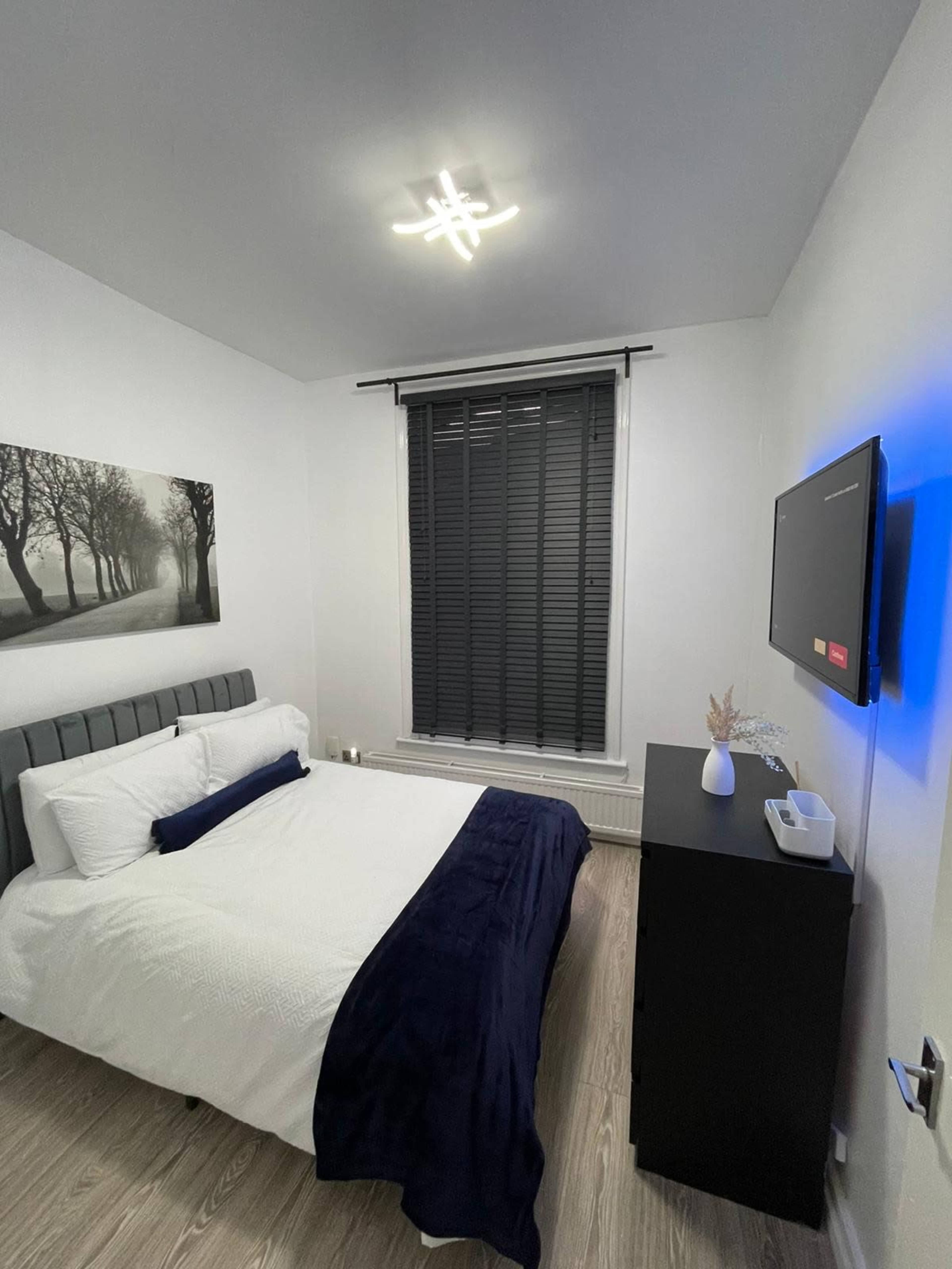 A small, modern bedroom featuring a bed with white bedding and a navy throw, a dresser, and a framed black-and-white landscape photograph on the wall.