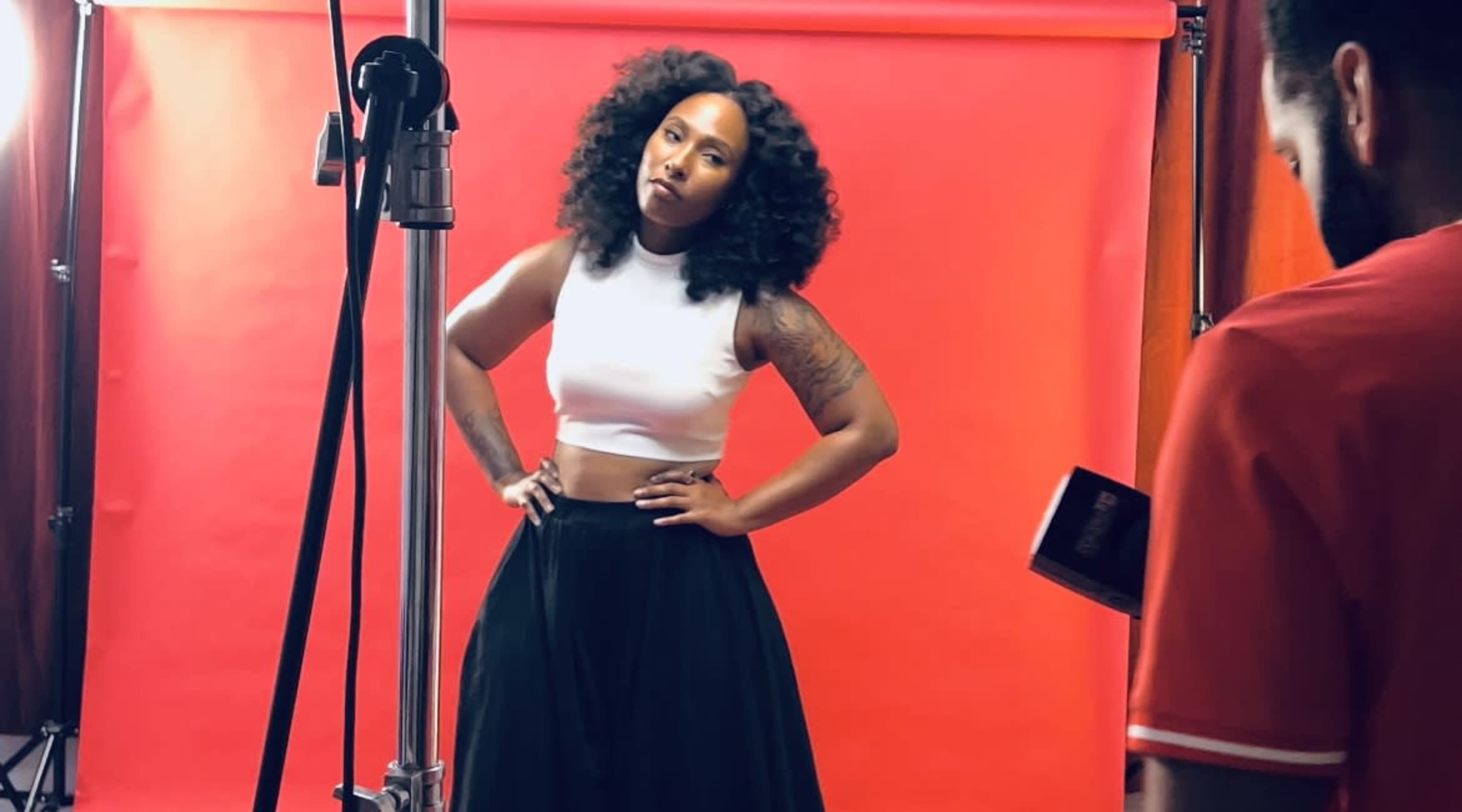 A woman poses with her hands on her hips in front of a red background during a photoshoot.
