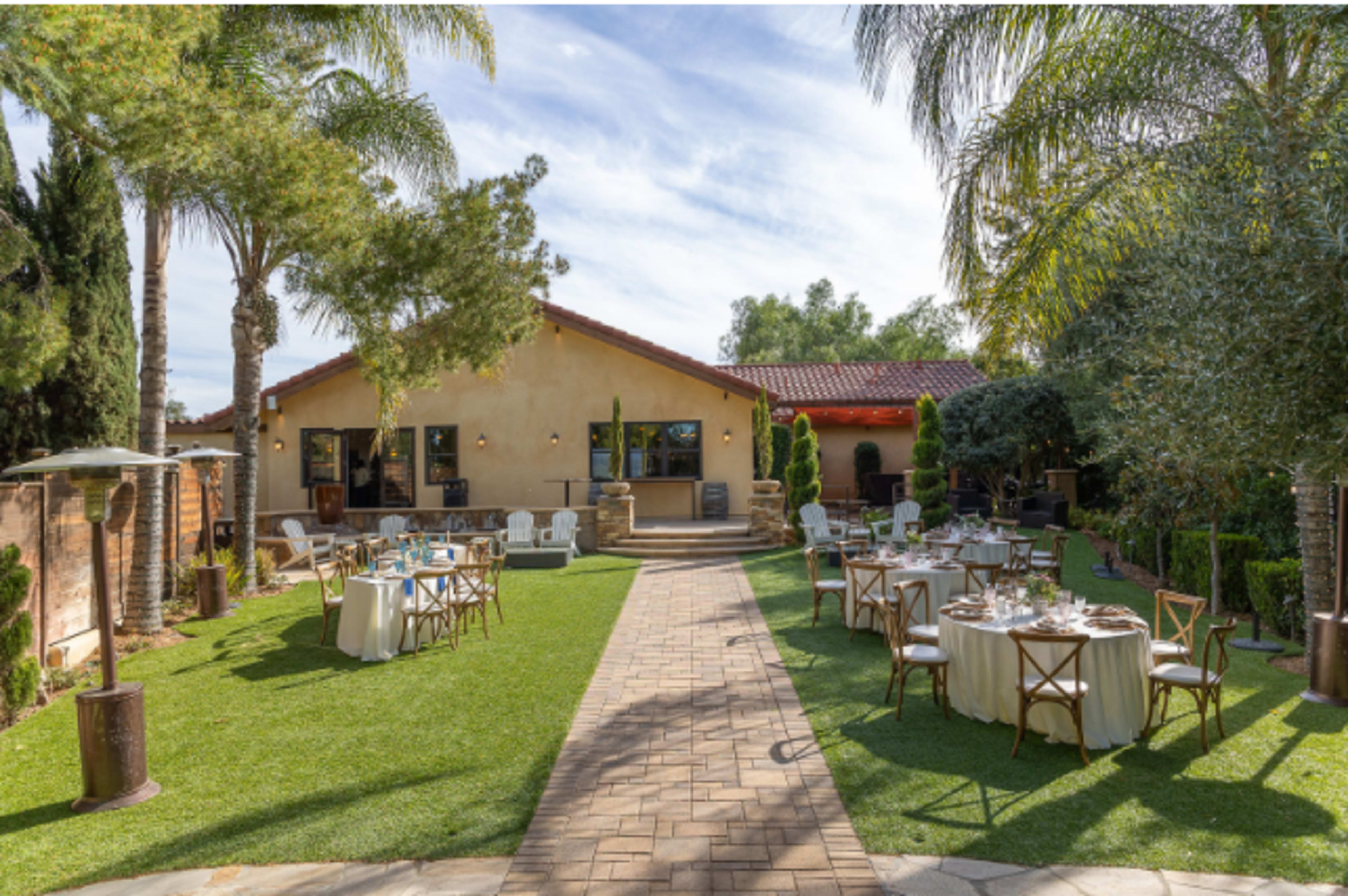 The image shows an outdoor garden setting with neatly arranged dining tables and chairs on a green lawn, surrounded by trees and a rustic building in the background.