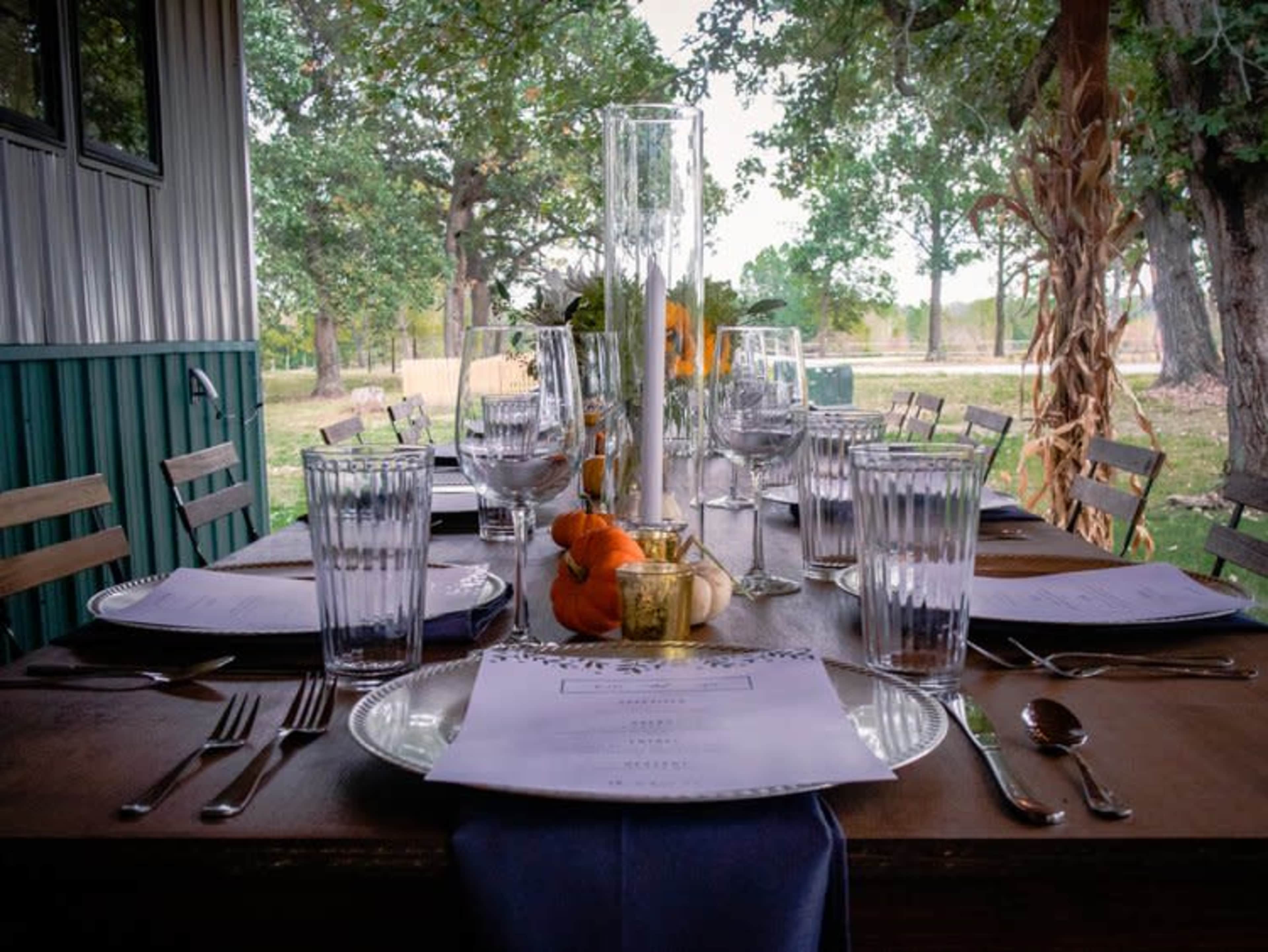 A dining table is set outdoors with glassware, plates, and menus, surrounded by trees and natural scenery.