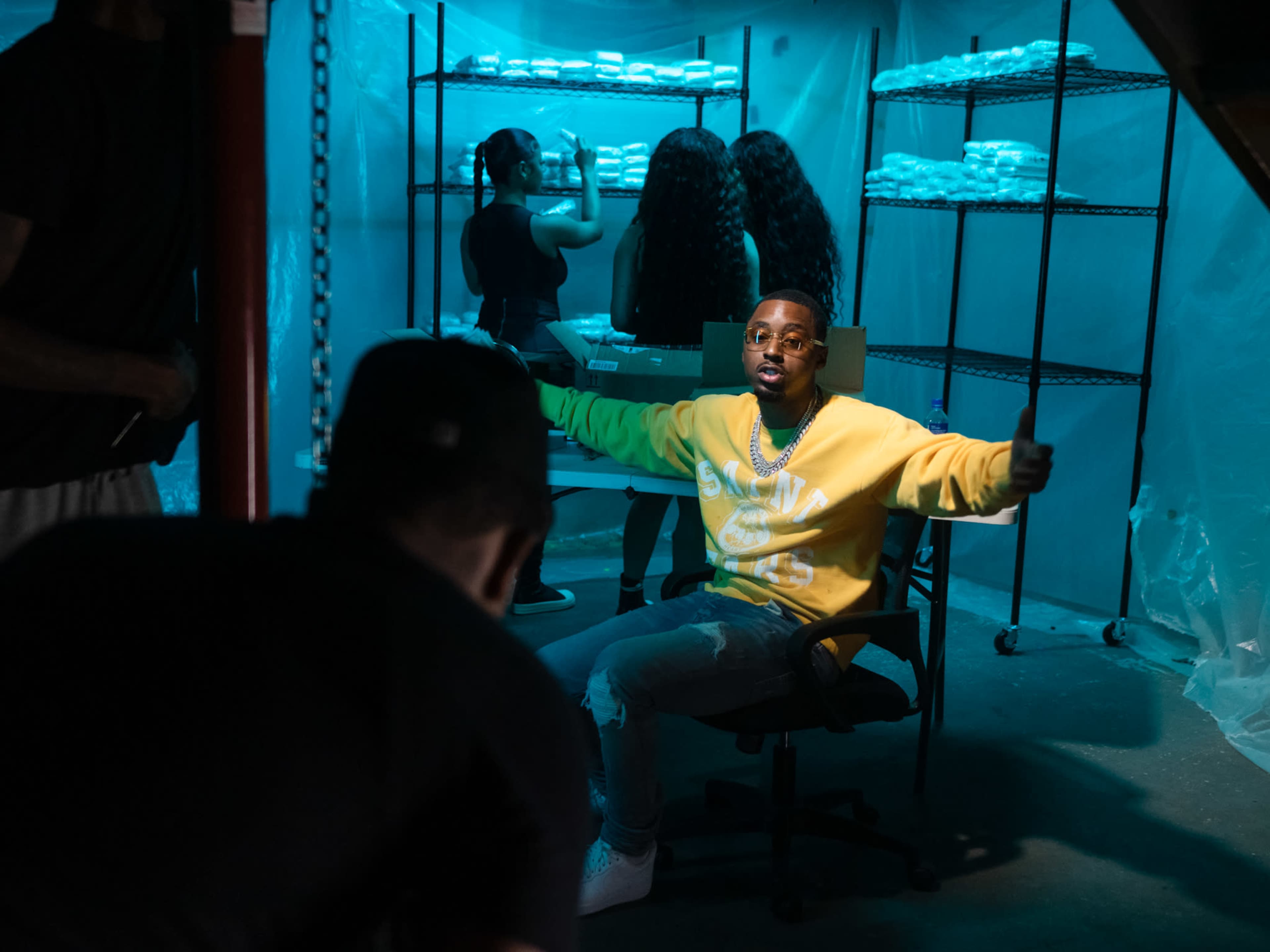 A person in a yellow sweatshirt sits in a chair in the foreground while two individuals work at a table behind them in a dimly lit, plastic-covered space.