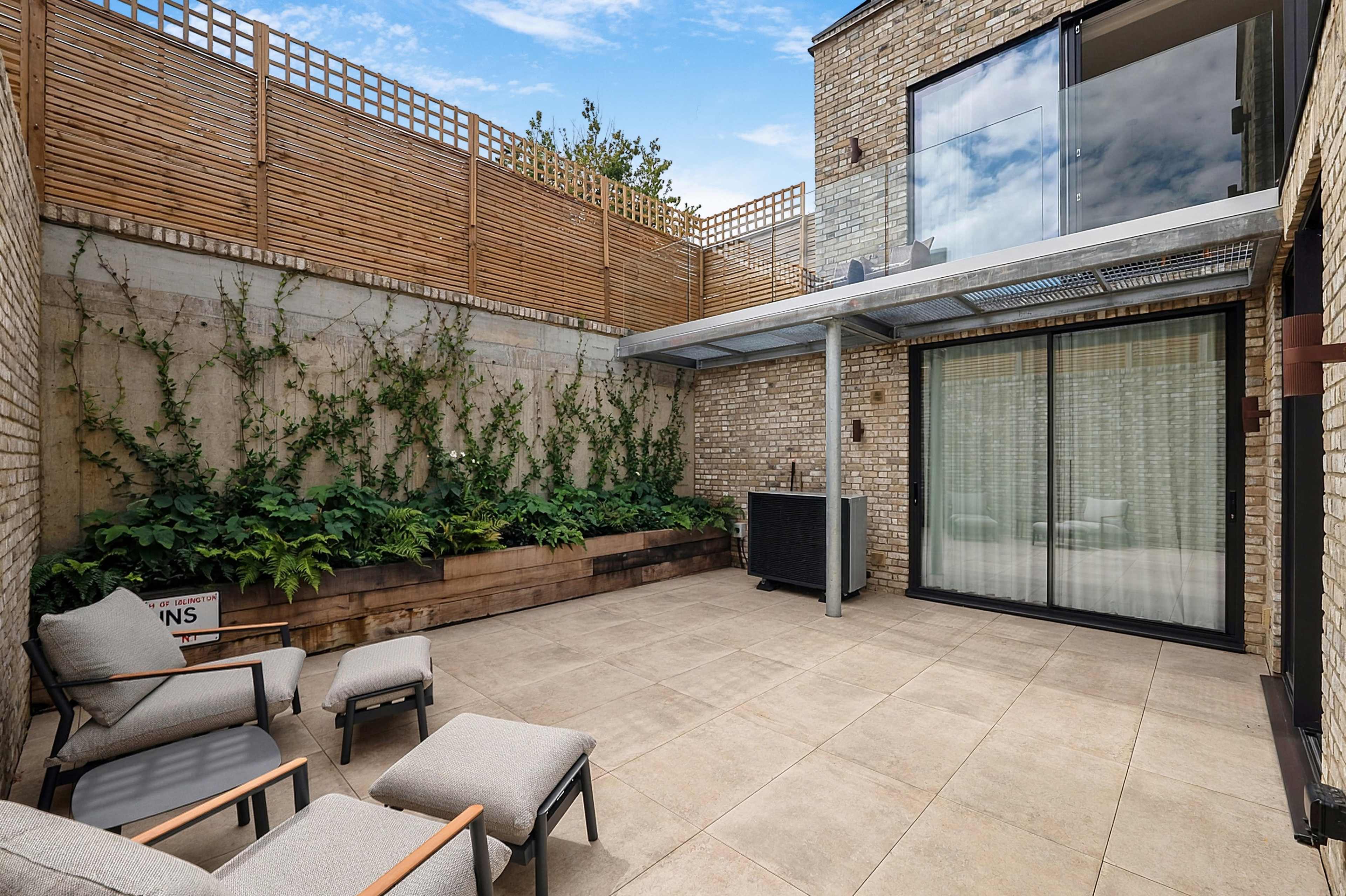 The image shows a modern outdoor patio surrounded by brick walls and a wooden fence, featuring seating and a planted area.