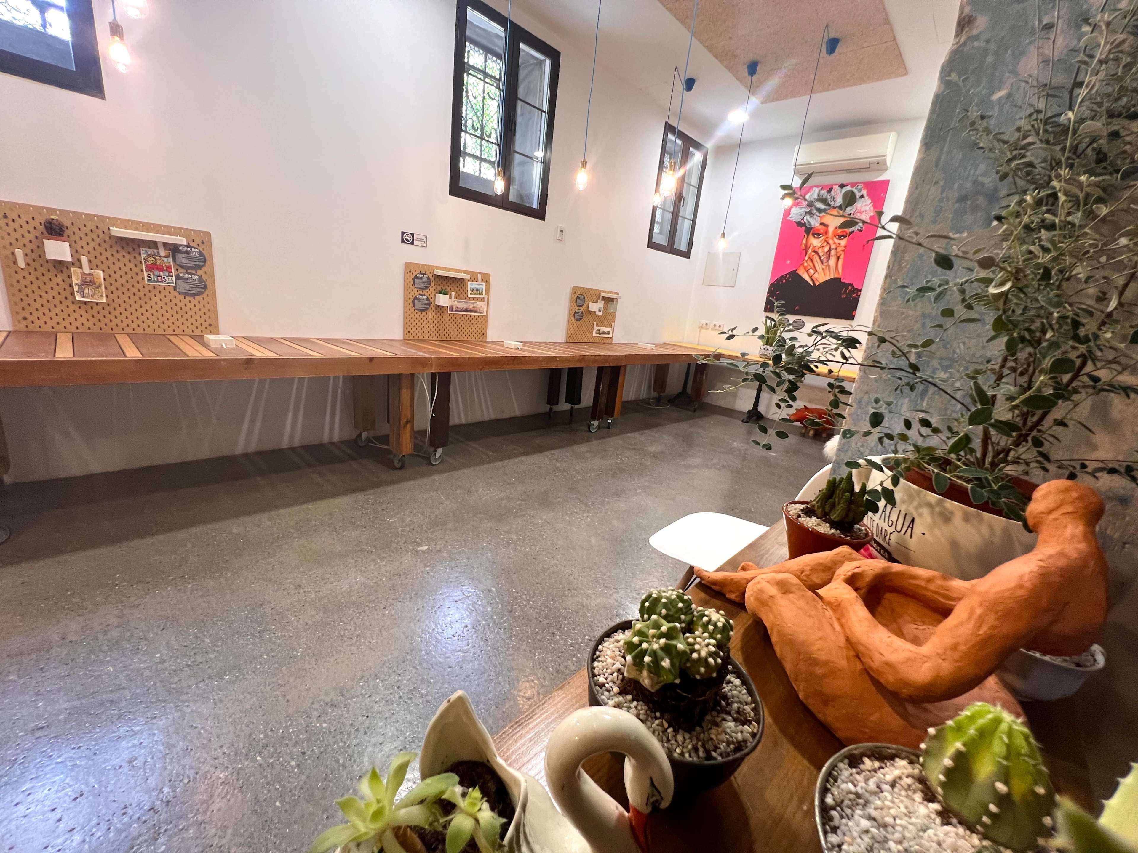 The image shows a modern workspace featuring long wooden tables, potted plants, and artistic elements in a well-lit room.