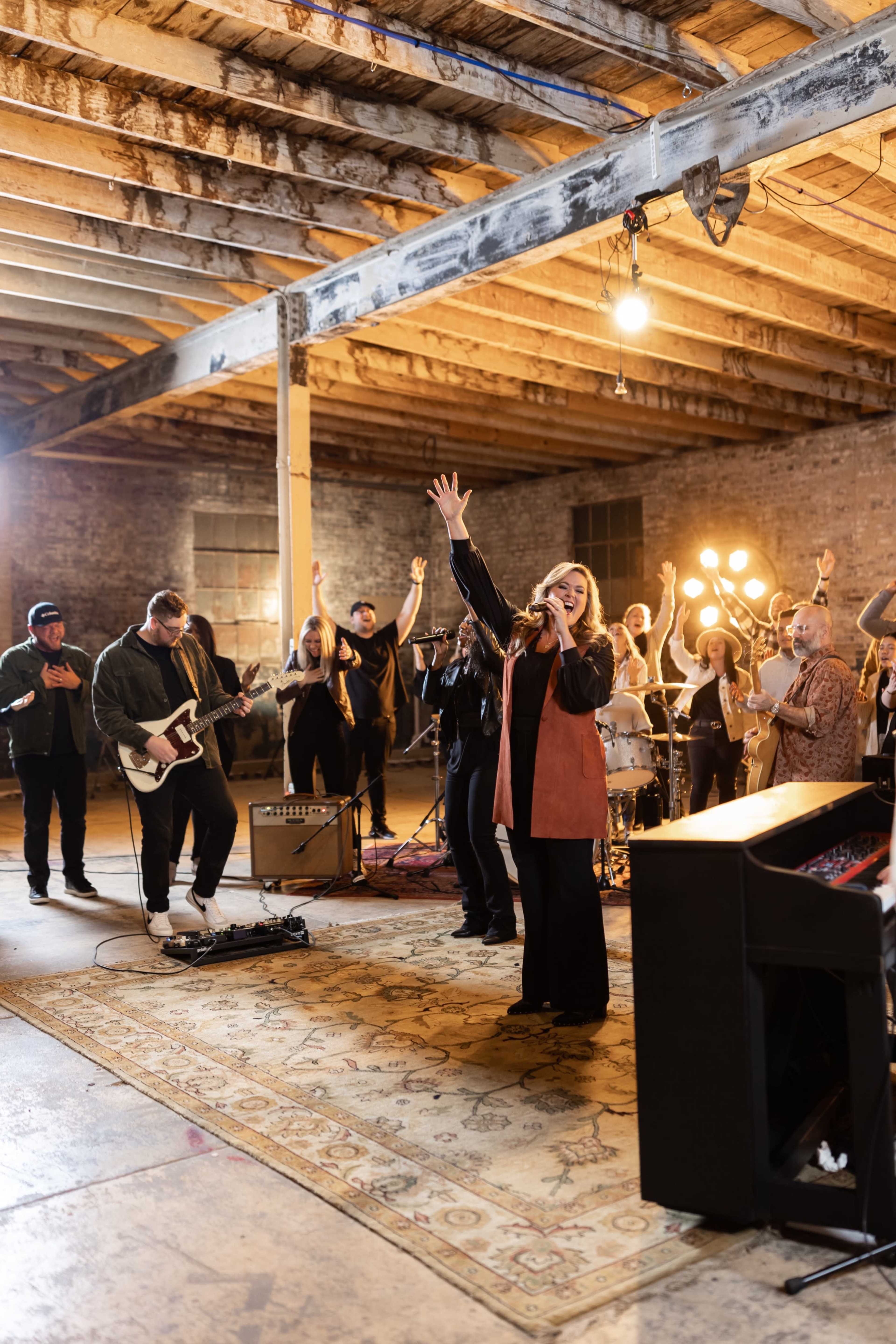 A musical group performs in a spacious, dimly lit warehouse, with a singer on stage and a diverse audience participating enthusiastically.