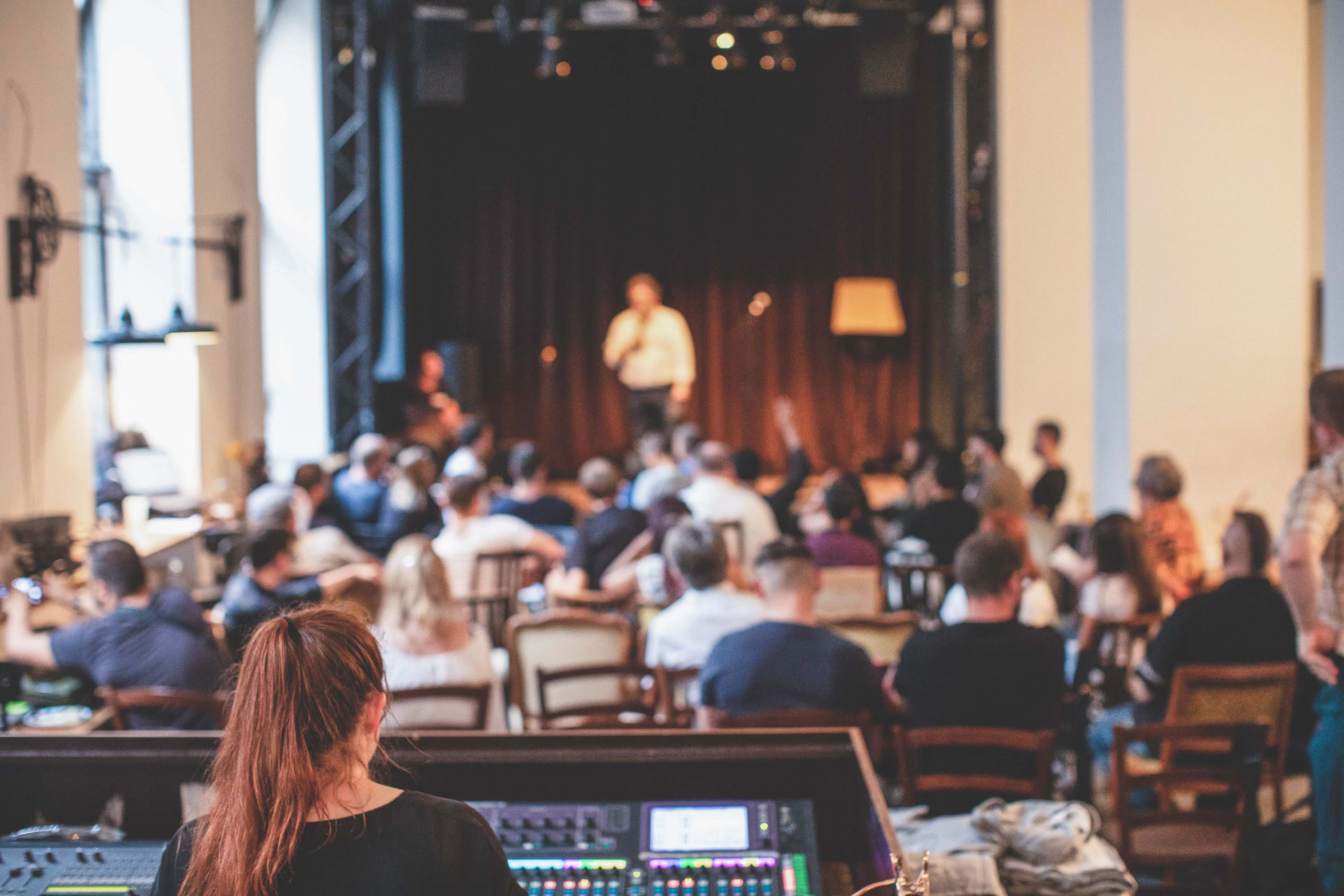 A crowd of people sits in a dimly lit venue, watching a performer on stage while a sound technician adjusts equipment in the foreground.