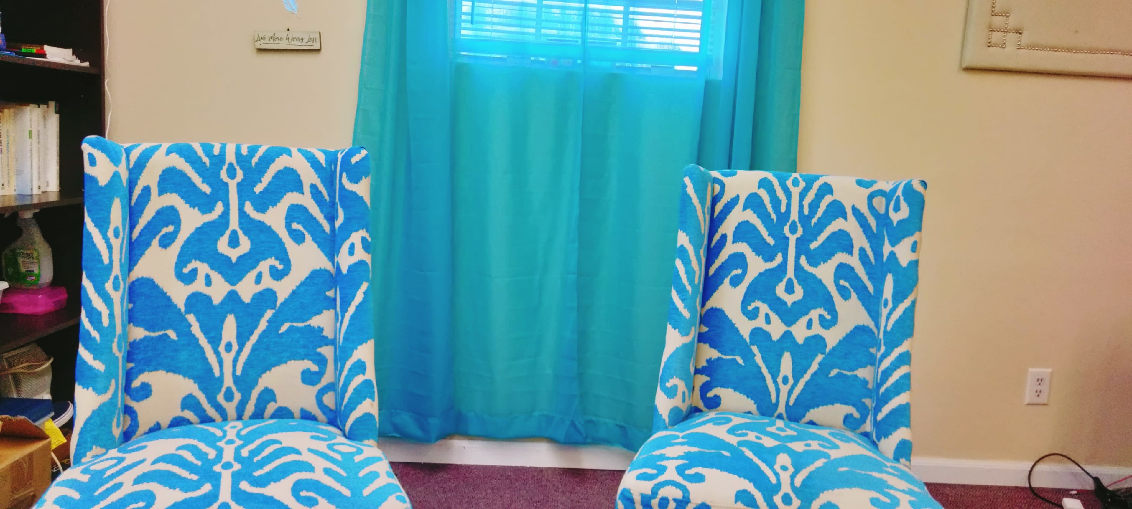 Two blue-patterned chairs face a window covered with a turquoise curtain in a brightly lit room.