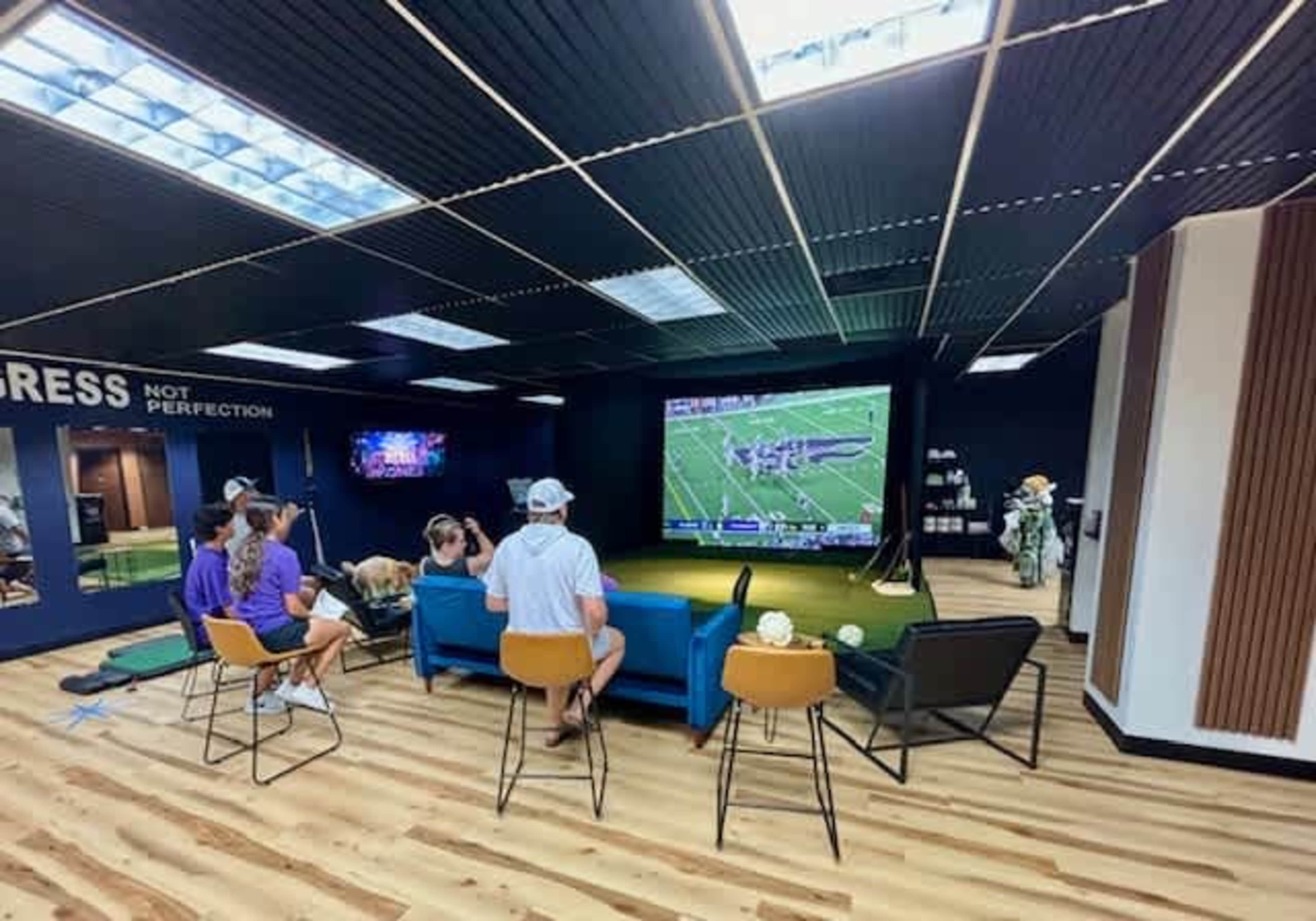 A group of people is seated on couches in a modern room, watching a football game on a large screen while surrounded by artificial grass and decorative plants.
