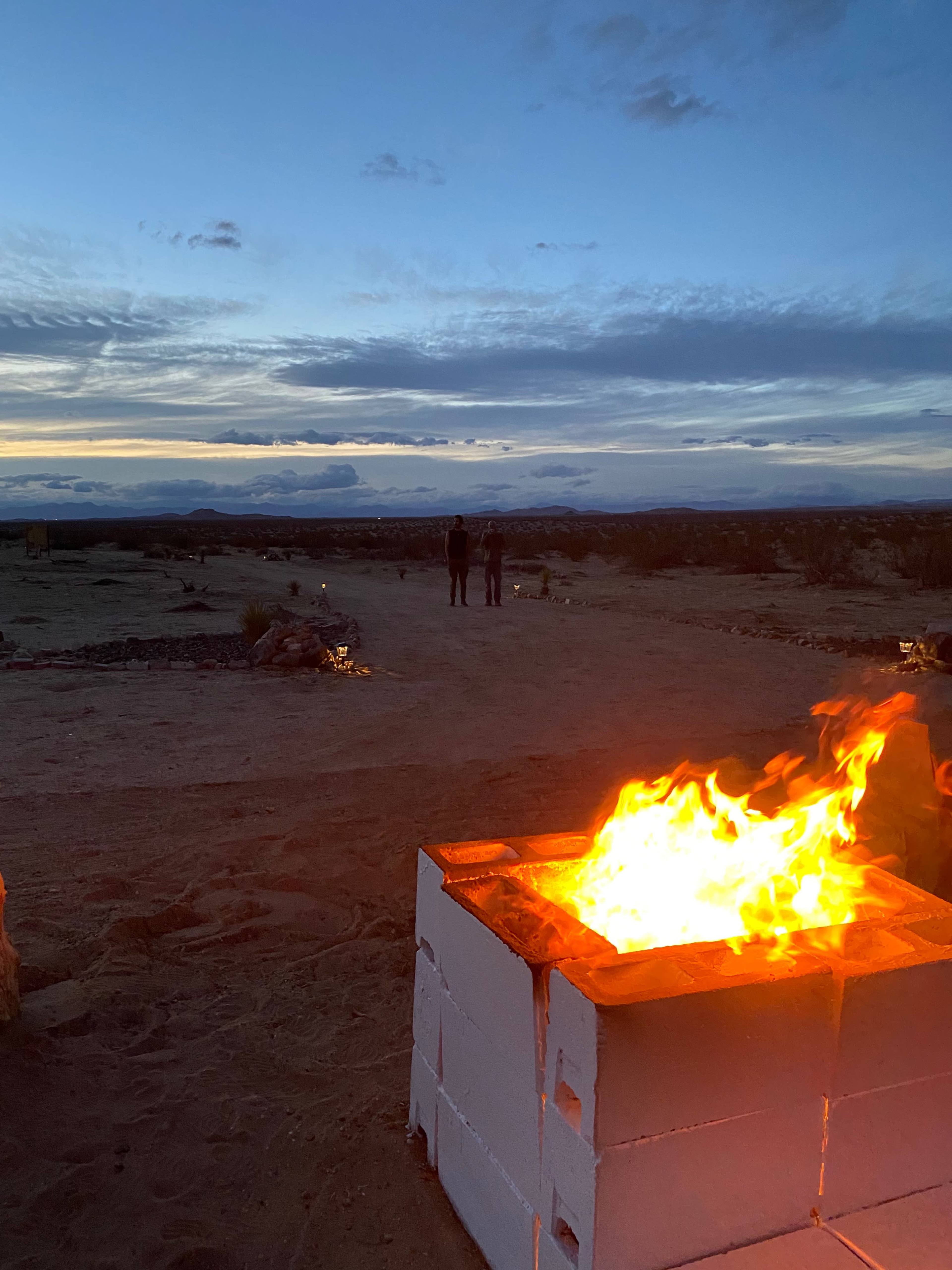 Two people stand in the distance near a fire pit as the sun sets over a desert landscape.