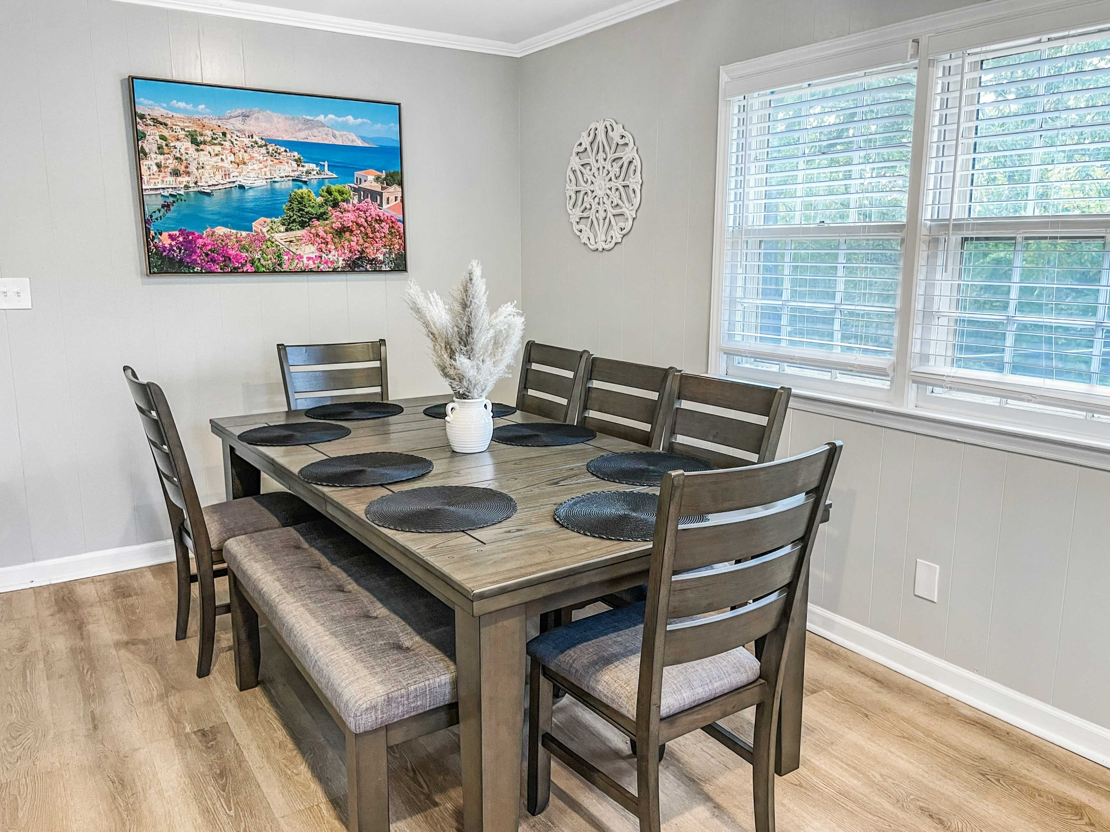 A wooden dining table with matching chairs is set in a bright room featuring a framed landscape picture on the wall.