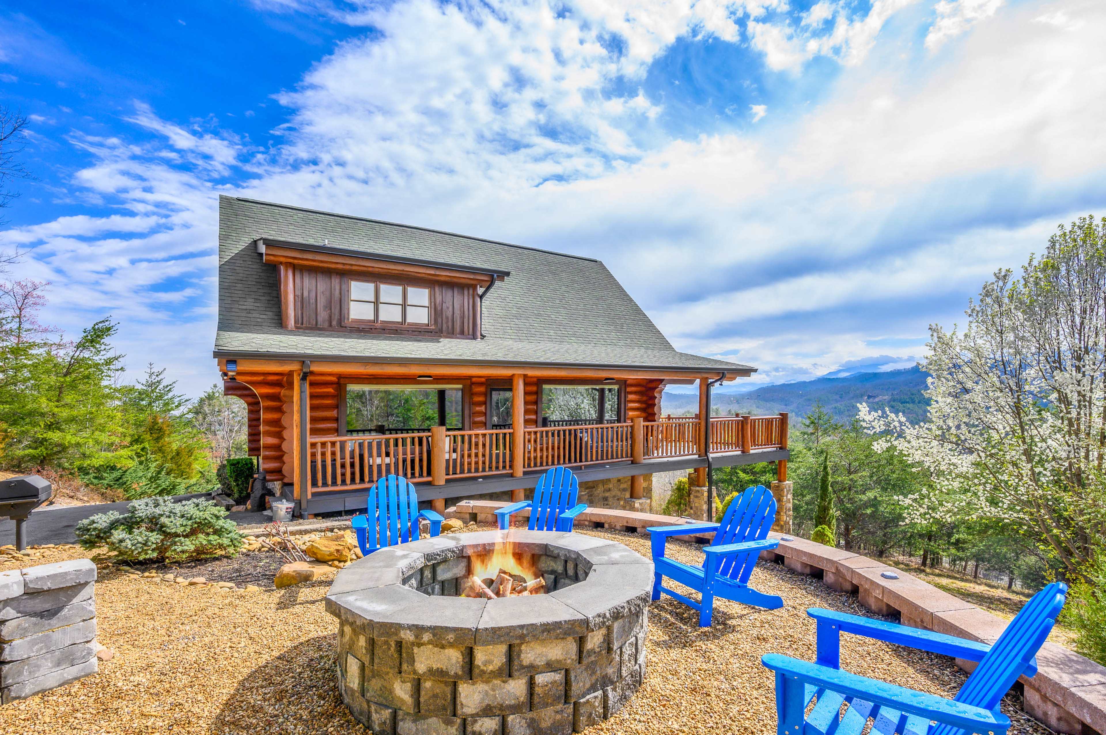A log cabin with a spacious porch overlooks a fire pit surrounded by blue Adirondack chairs and a scenic landscape.