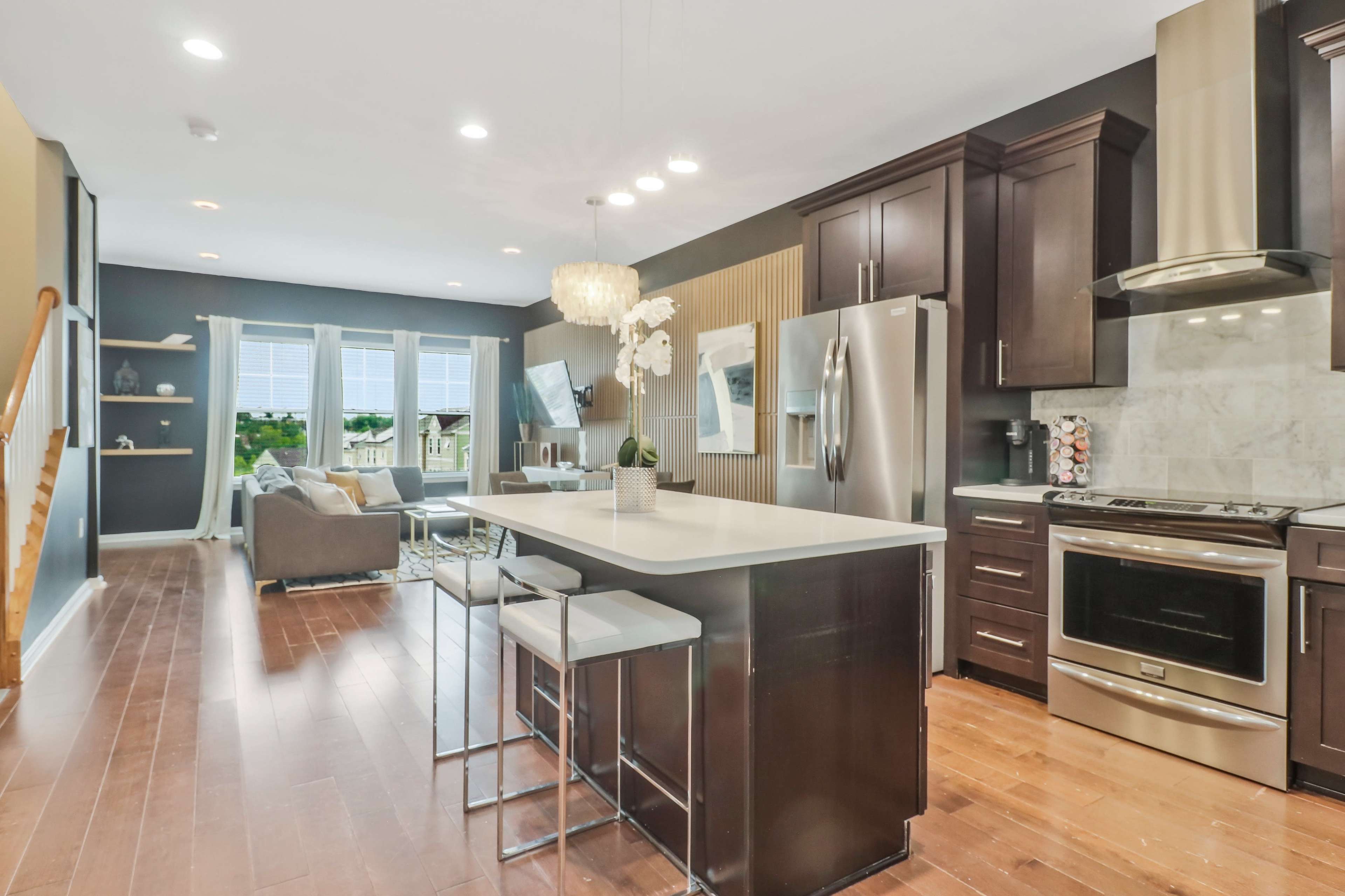 A modern kitchen features dark cabinets, stainless steel appliances, and a large island with seating, adjacent to a bright living area with large windows.
