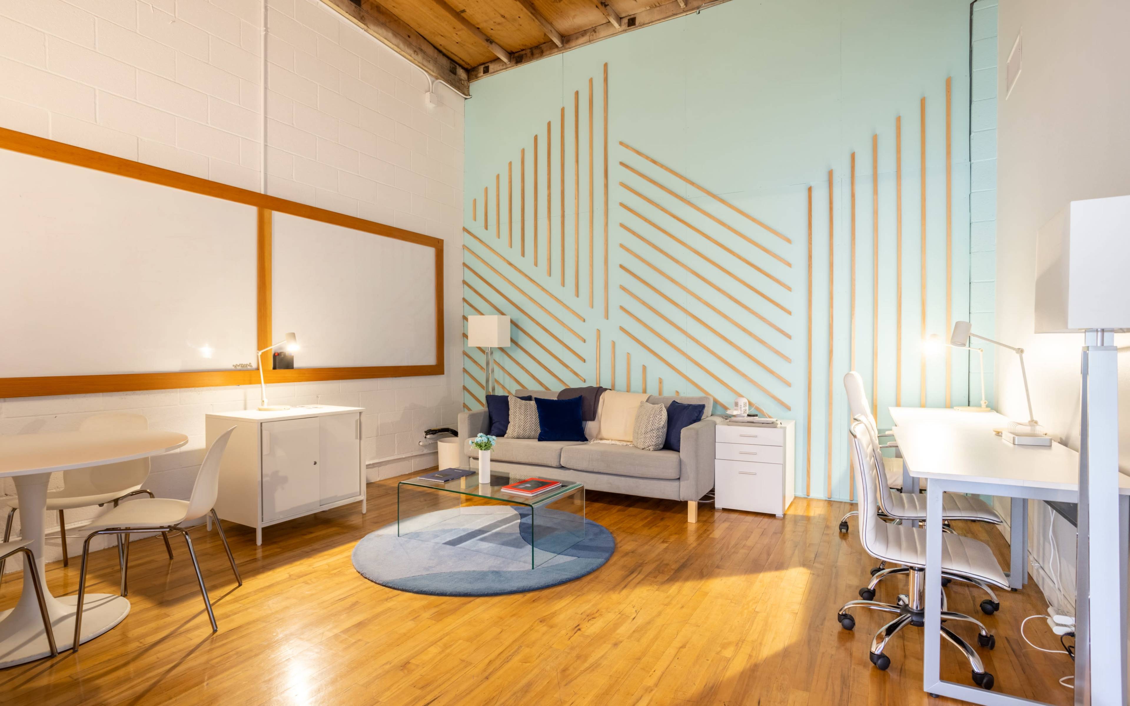 A modern workspace featuring a gray sofa, white desks with chairs, a round rug, and a textured wall accentuated with wooden stripes.